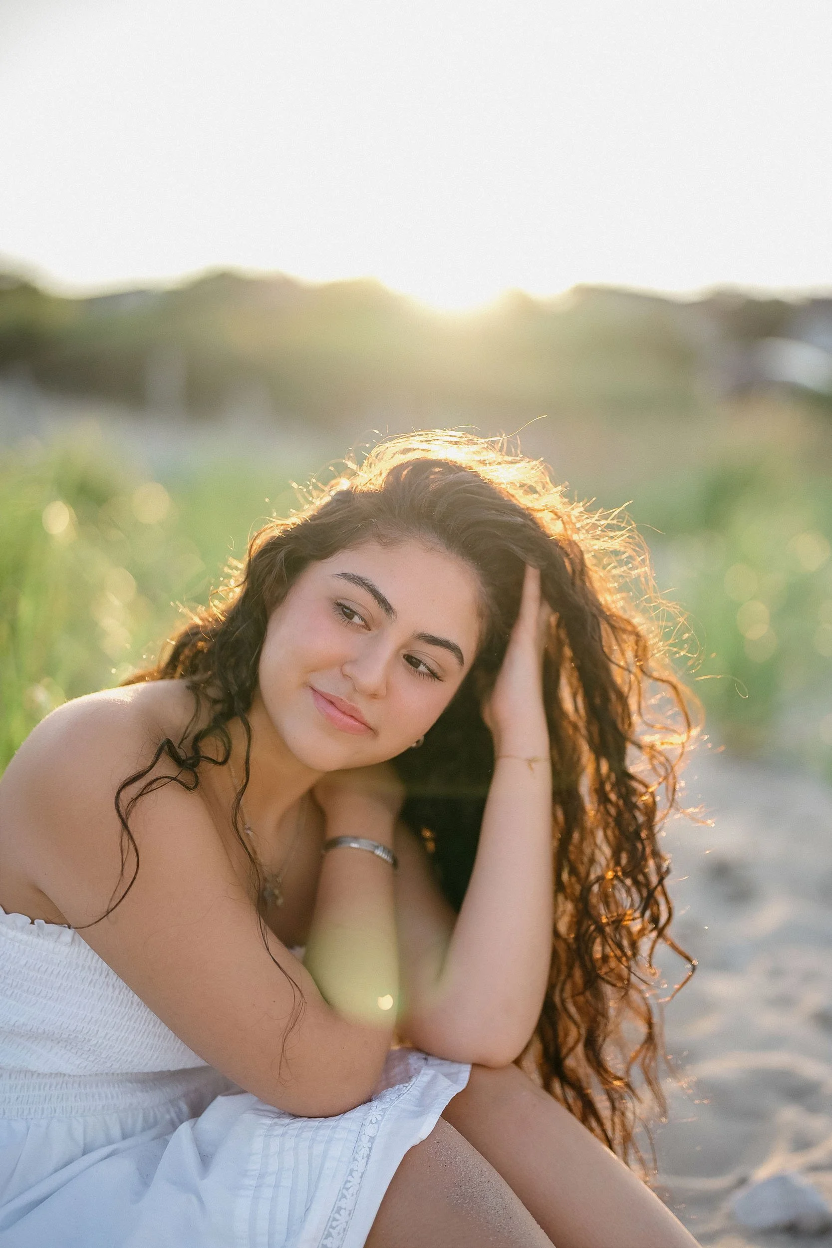 A young woman with long curly hair sitting on a sandy beach at sunset, wearing a white dress, with her hand resting on her head and looking thoughtfully to the side.