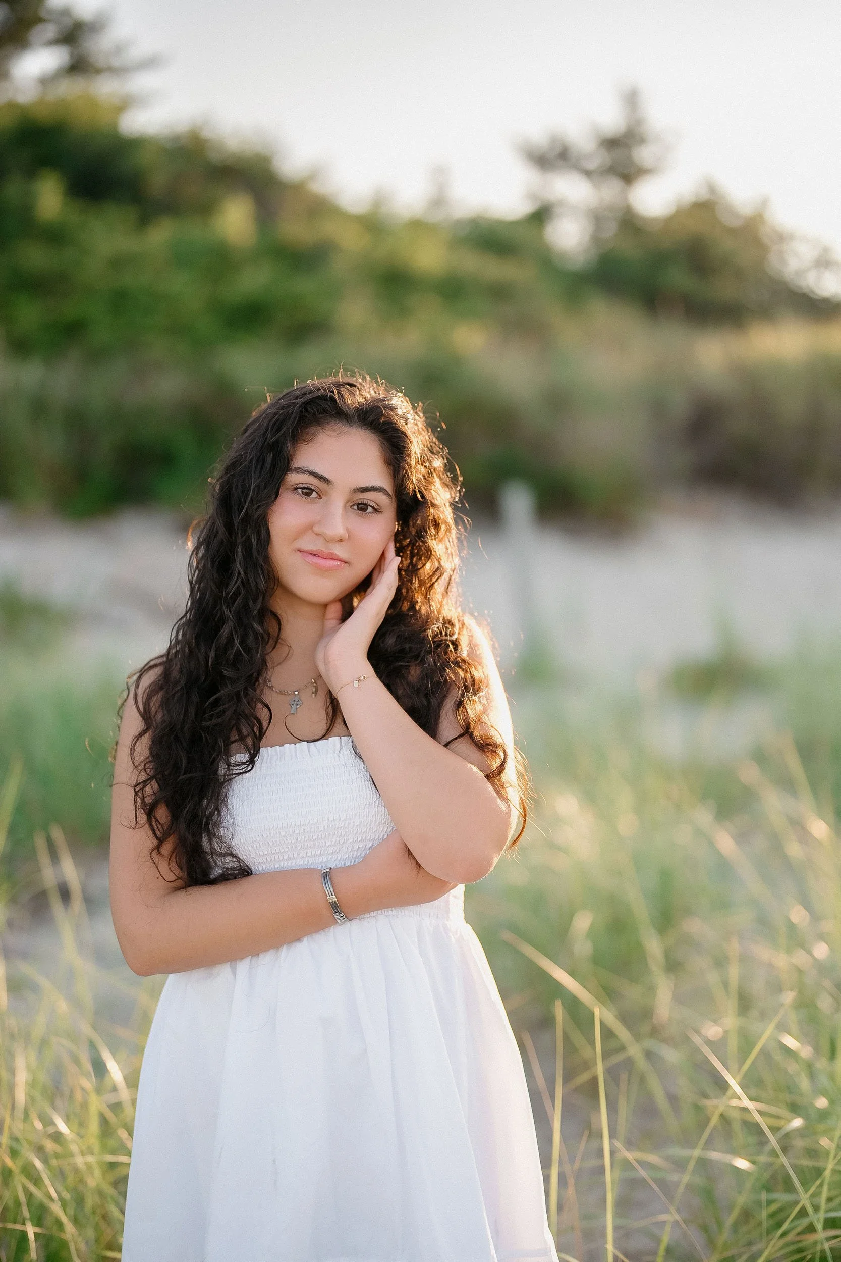 Young woman with long curly dark hair wearing a white dress, standing outdoors in a grassy area, with trees and a light sky in the background, during golden hour.