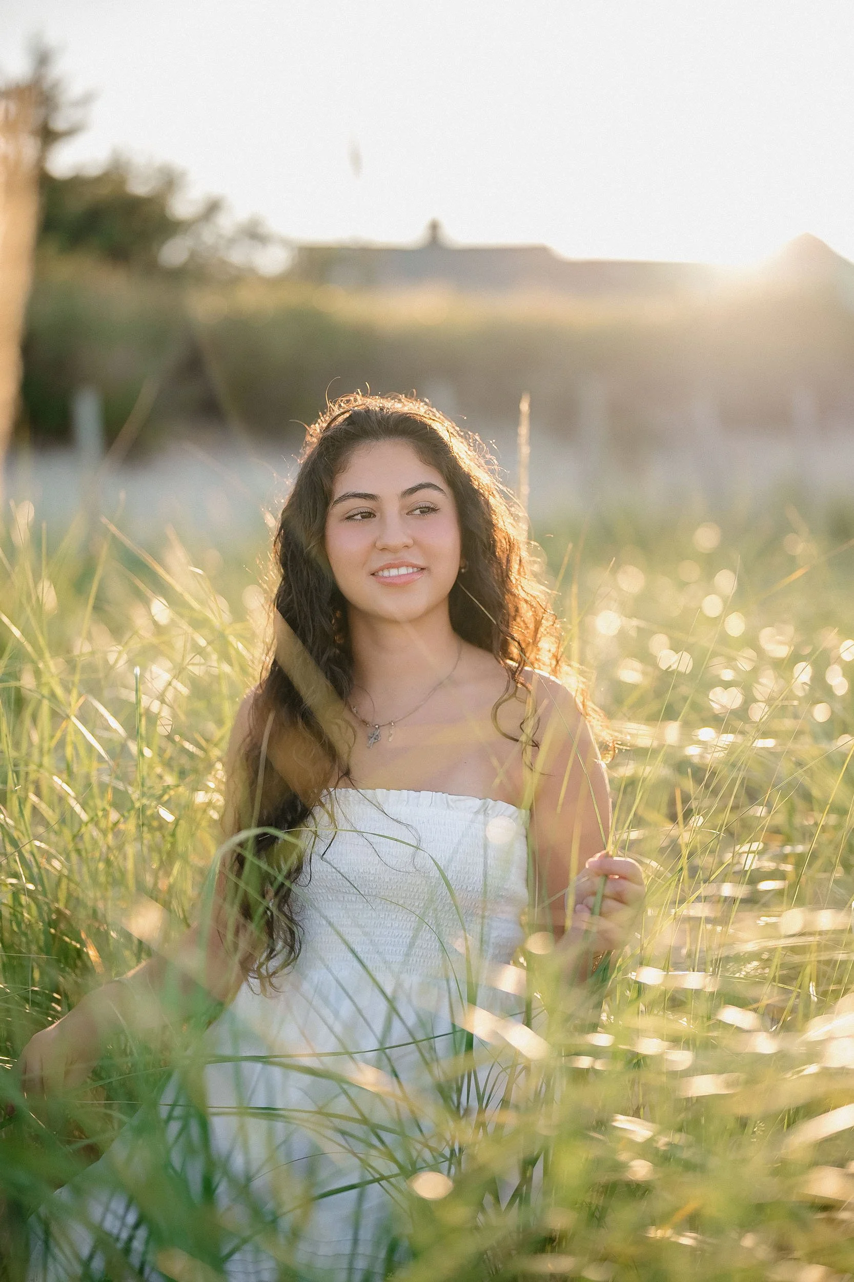 Young woman with long, curly hair standing in a field of tall grass, smiling slightly, with the sun setting behind her.