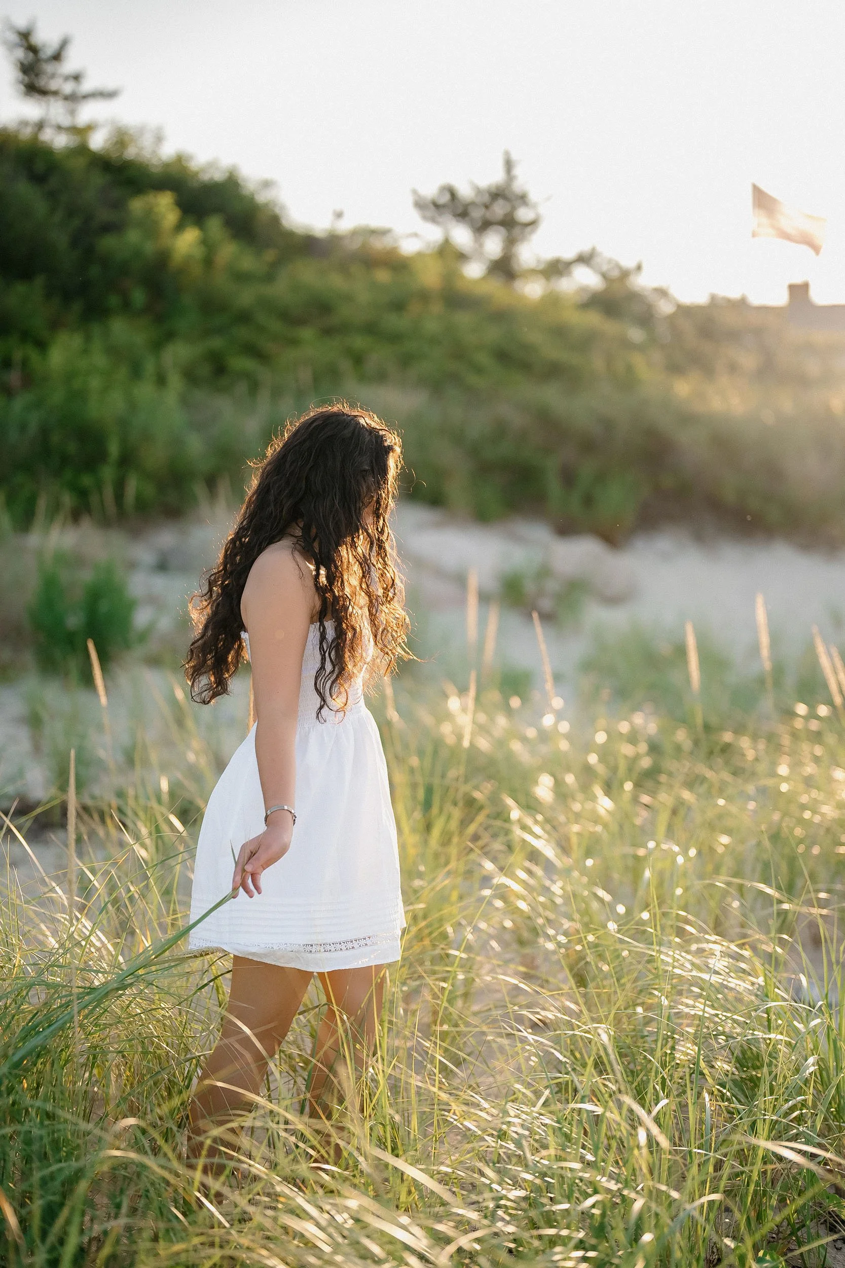 A woman with long curly hair wearing a white dress standing in tall grass on a beach at sunset.