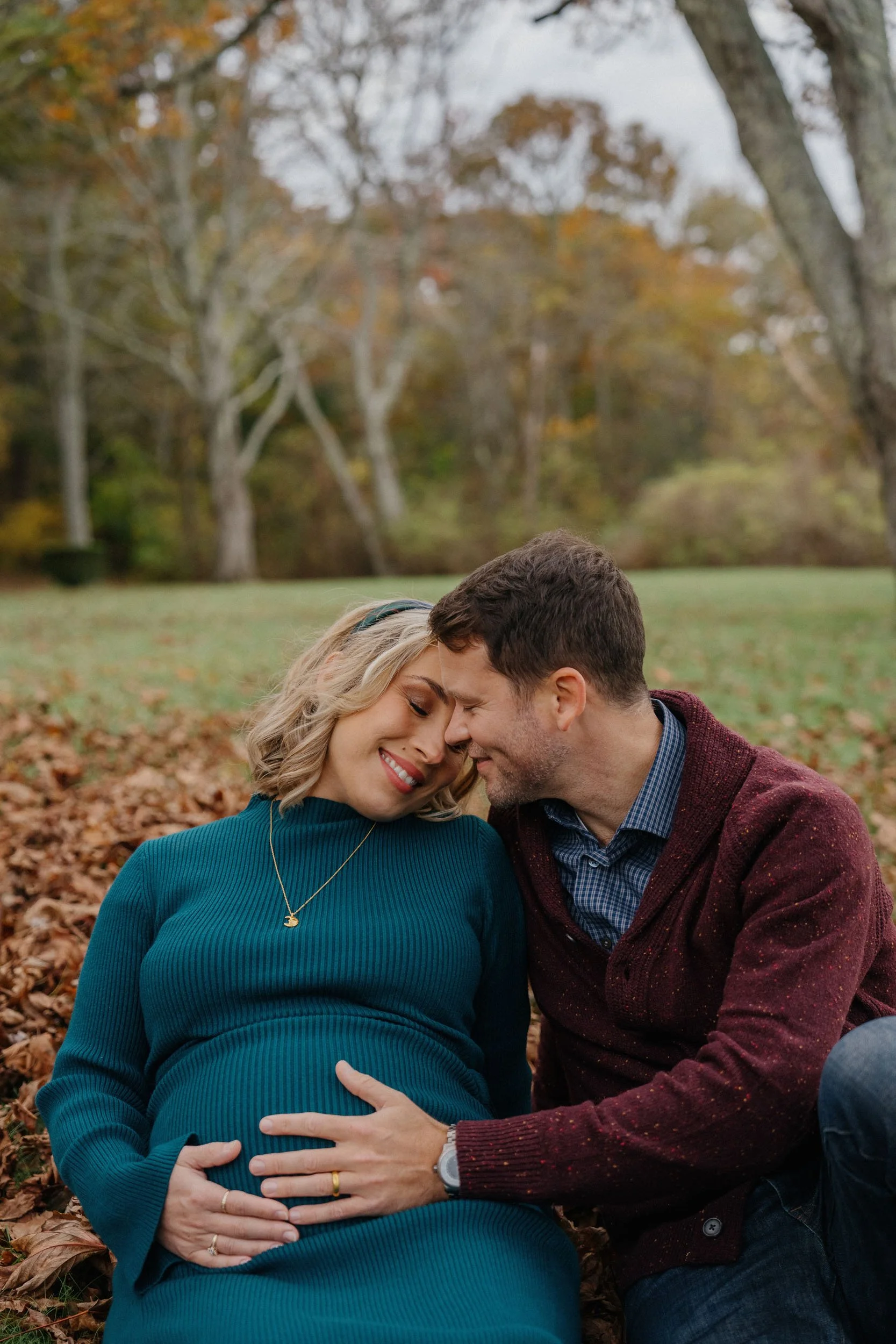 A pregnant woman and a man sitting on autumn leaves in a park, touching foreheads and smiling.
