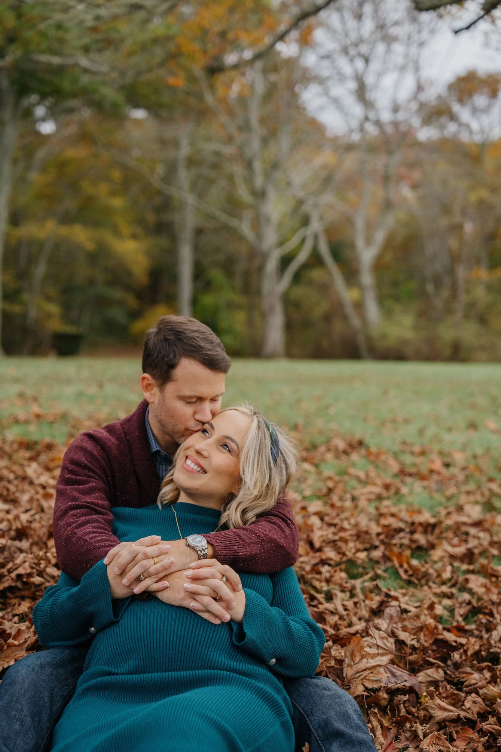 A couple sits on autumn leaves in a park, with the man kissing the woman's forehead and the woman smiling, surrounded by trees with colorful fall foliage.