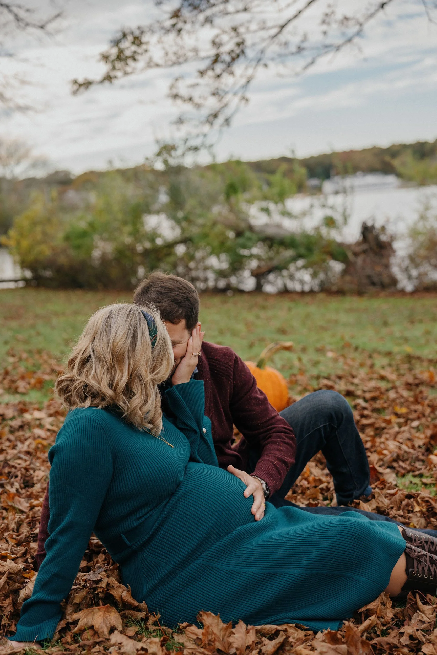 A couple sitting on fallen autumn leaves near a lake, with a large pumpkin behind them, during daytime. The woman, who appears pregnant, is touching her belly while the man hides his face in her hand.