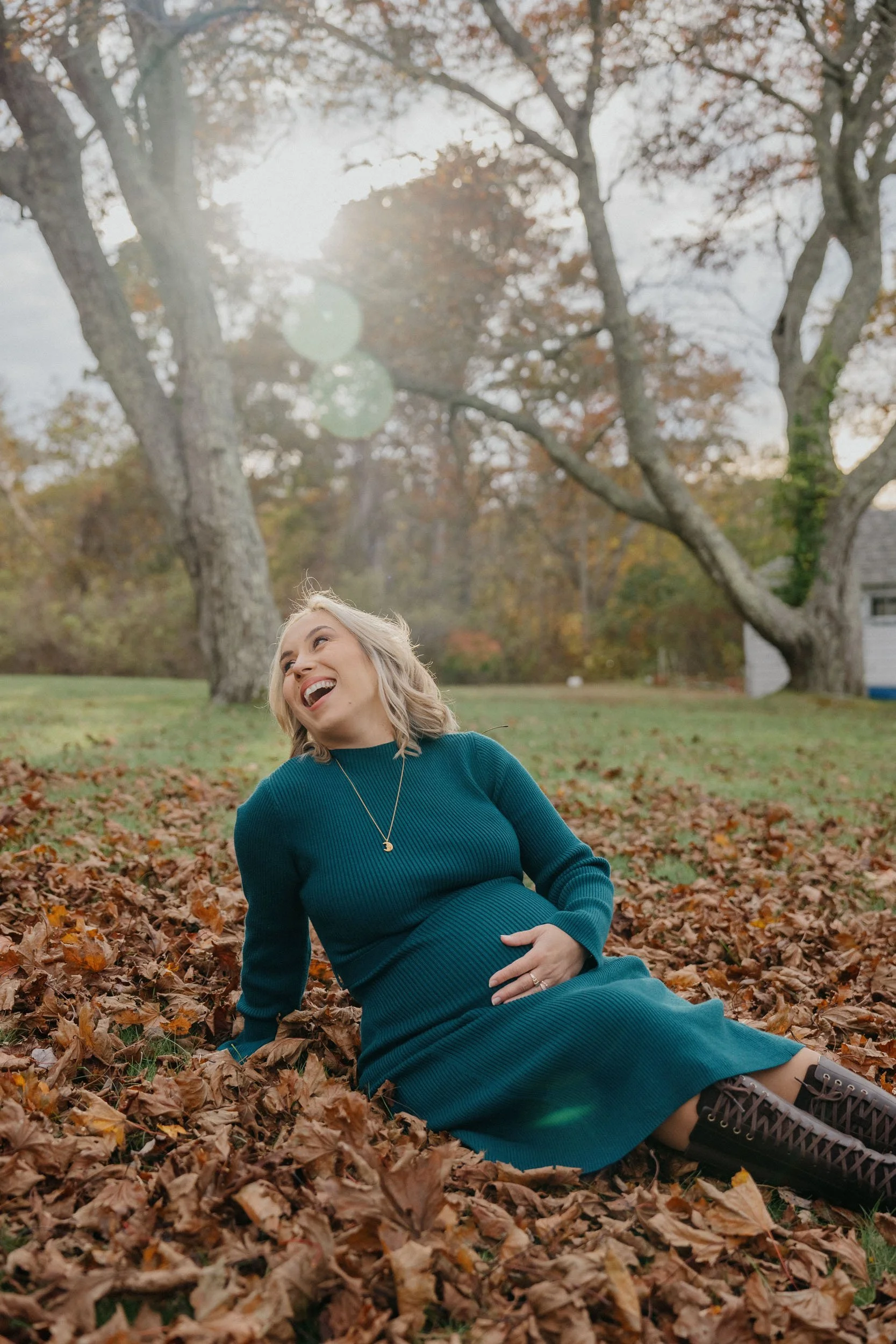 A pregnant woman with blonde hair wearing a teal dress and black boots, sitting on fallen autumn leaves in a park with trees in the background. She is laughing and holding her belly.