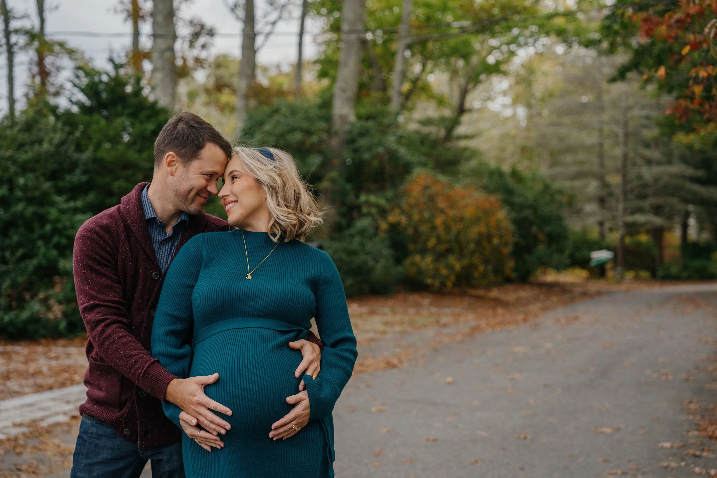 A couple, a pregnant woman and a man, stand close together outdoors during fall, touching her belly and smiling at each other.