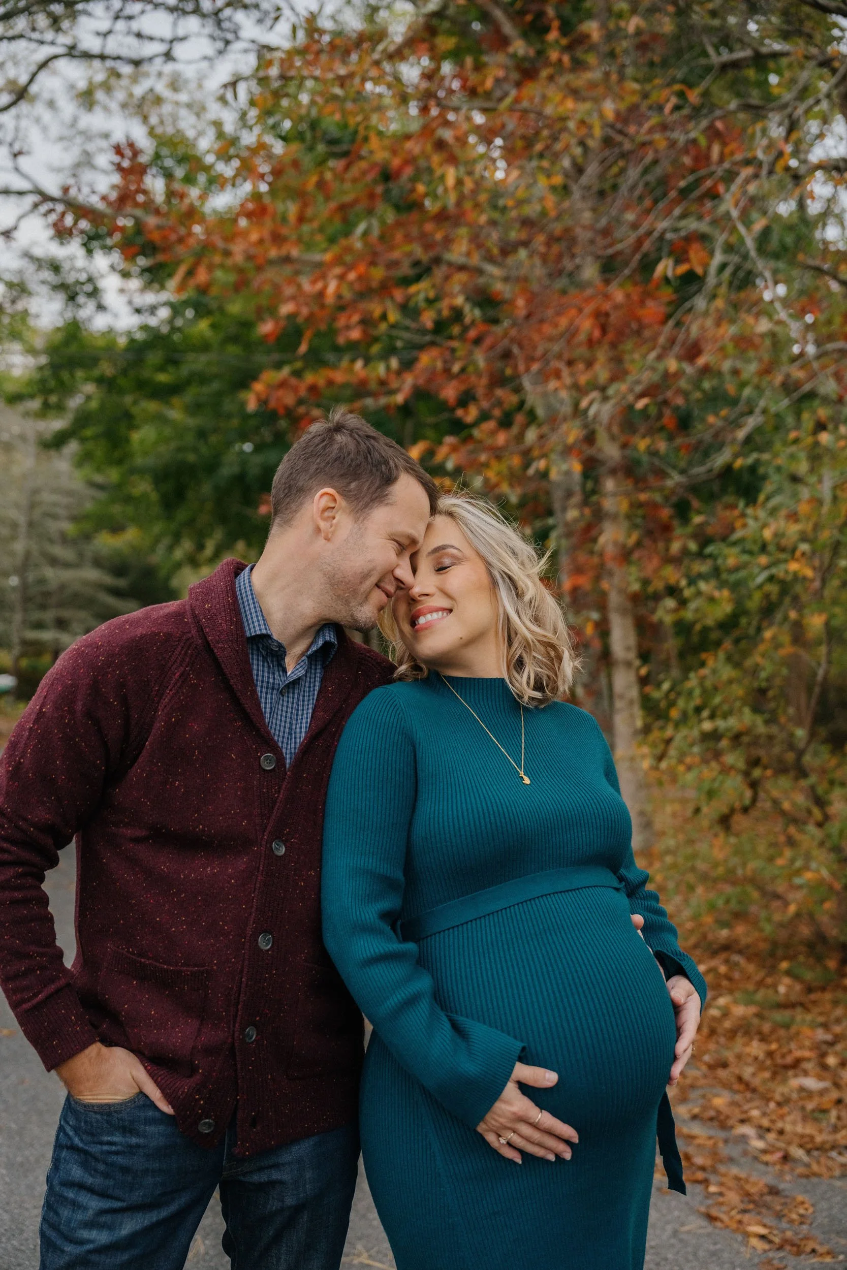 A couple sharing a tender moment outdoors during fall, with a background of colorful autumn trees. The woman is pregnant and gently holding her belly, smiling with her eyes closed as the man leans in close, their foreheads touching, both smiling.