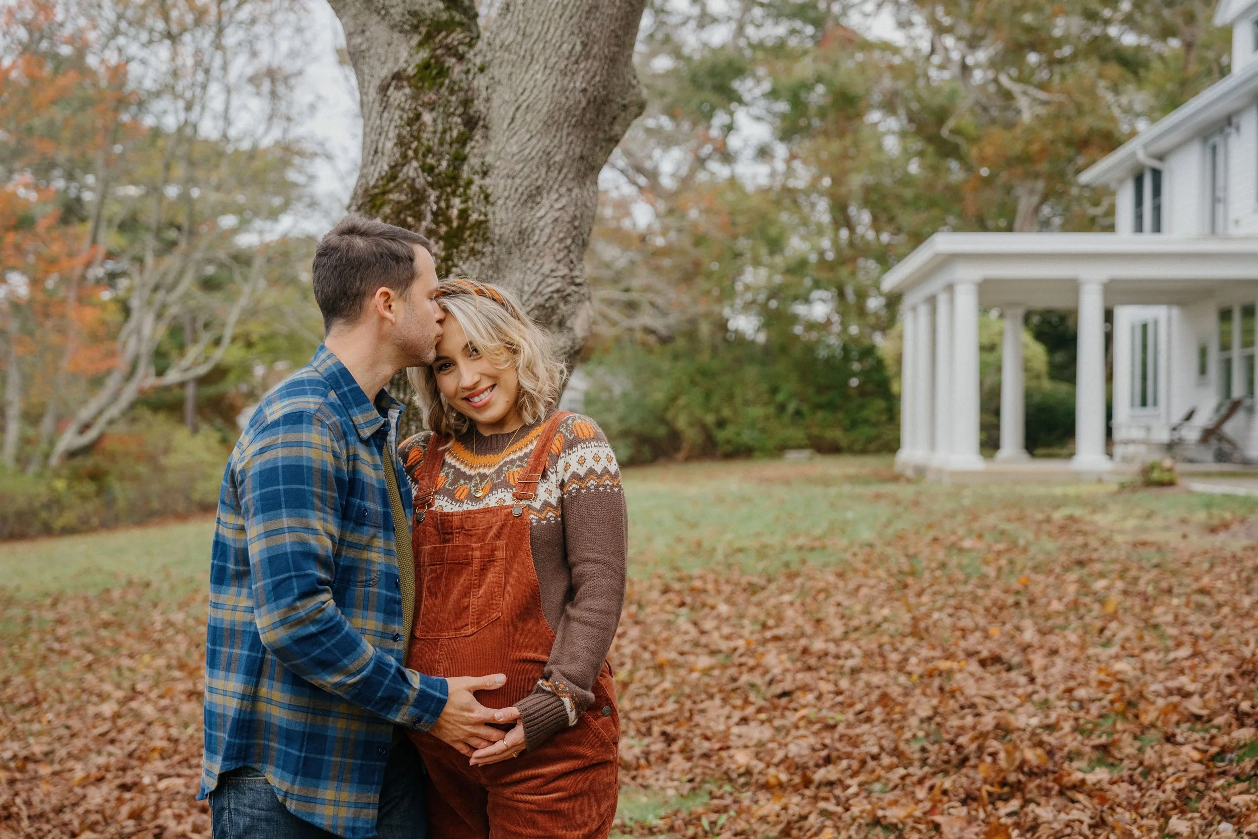 A couple standing outdoors in fall, with a young woman smiling and a man kissing her forehead, holding her pregnant belly, near a large tree, with a white house in the background.