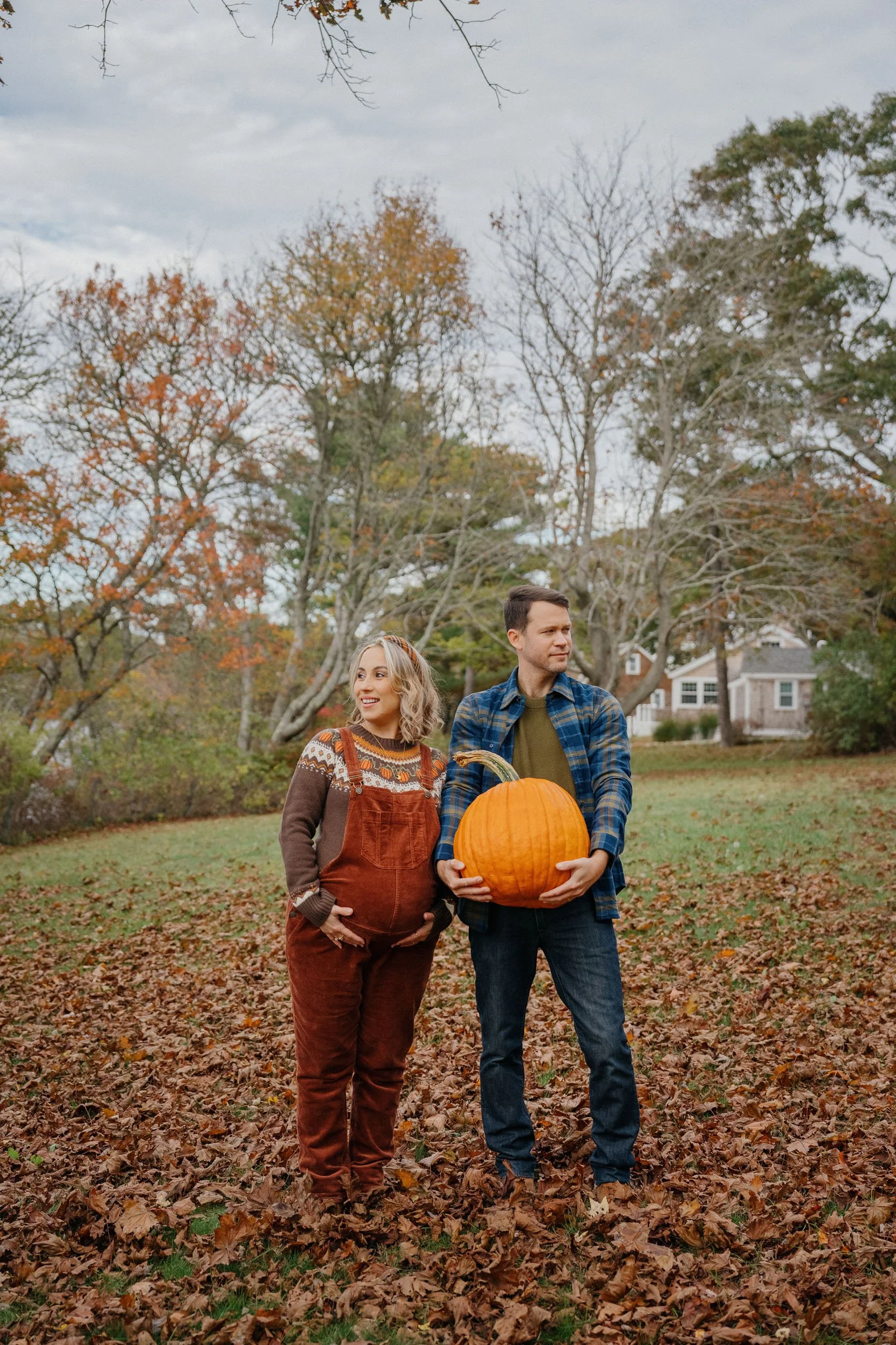 A couple standing outdoors in a park during autumn, holding a large pumpkin. The woman is pregnant and smiling, wearing brown overalls and a patterned sweater, while the man is looking to the side, dressed in a blue checkered shirt and jeans. The gro