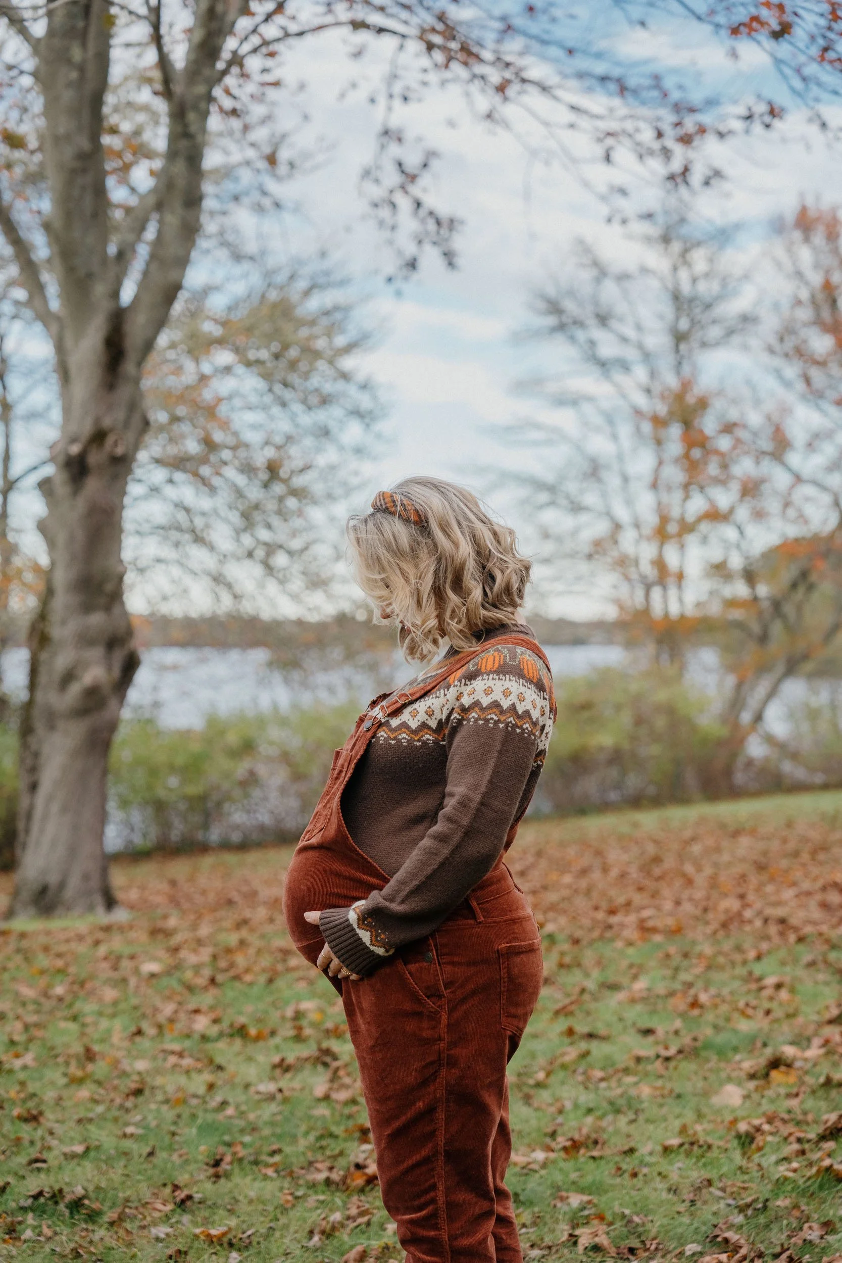 A pregnant woman with blonde hair wearing a patterned sweater and rust-colored overalls standing outdoors in a park with autumn trees and fallen leaves.