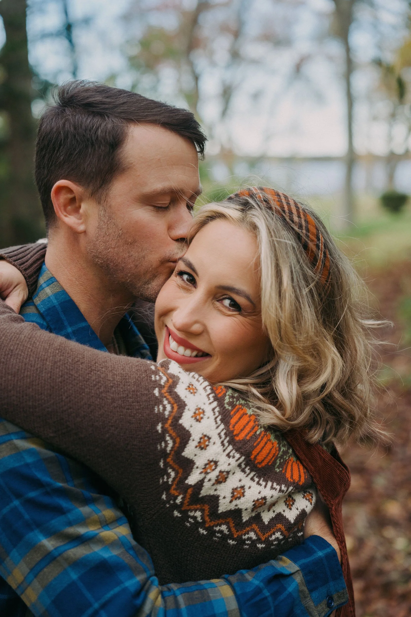 A man kisses a woman on the cheek while hugging her outdoors in a wooded area, both smiling happily.