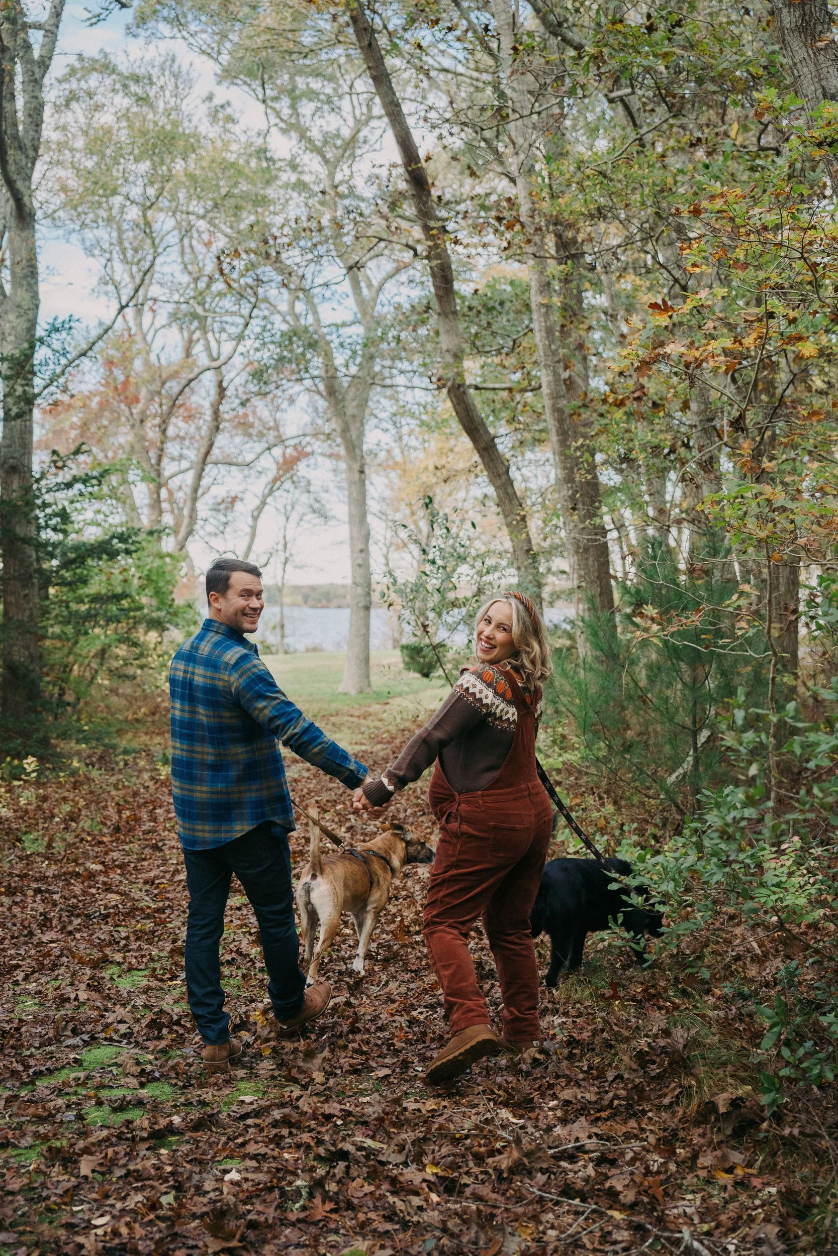 A smiling couple holding hands and walking through a fall forest trail with two dogs, one tan and one black, among trees with autumn leaves.