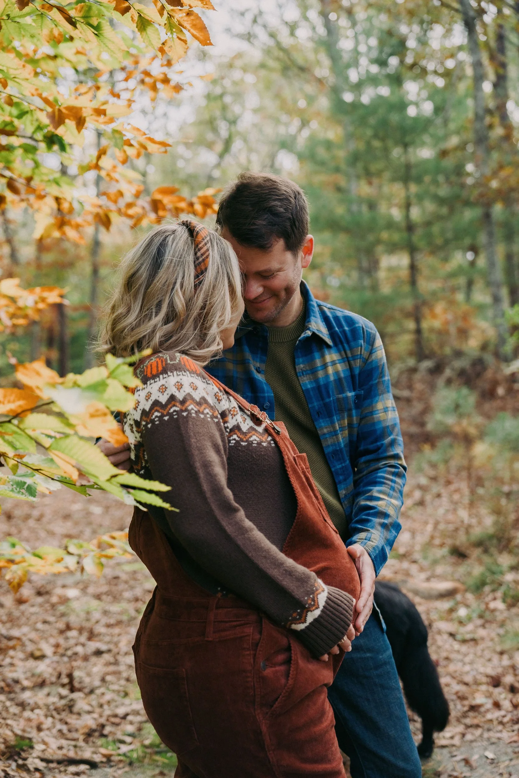 A couple standing close together in a forest during fall, smiling and touching foreheads. The woman wears a patterned sweater and overalls, the man wears a plaid shirt and jacket.