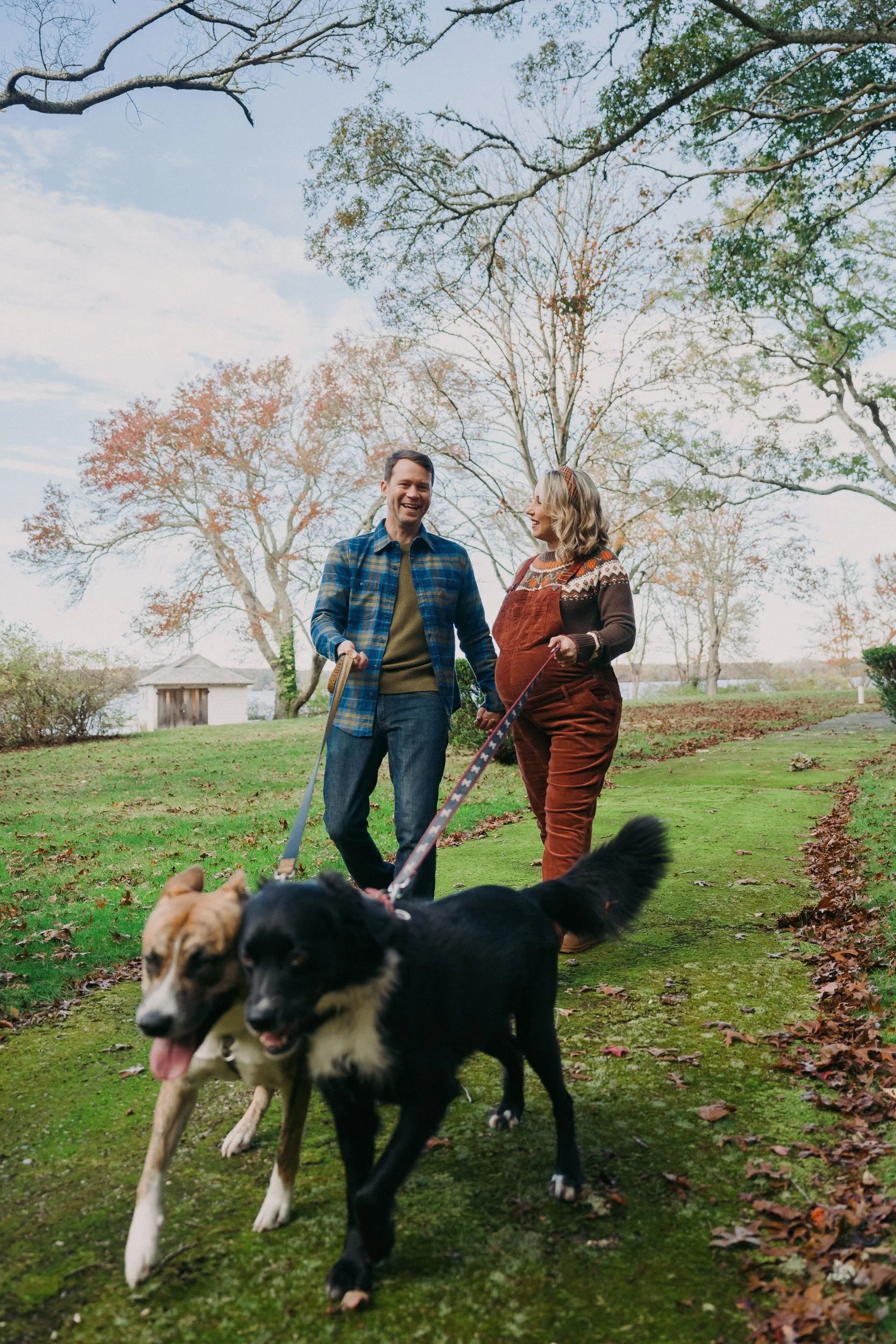 A couple walks with two dogs in a park during autumn. The man wears a blue plaid shirt and jeans, and the woman wears a brown jumpsuit with colorful pattern details. They are smiling and holding hands as they walk along a green, leaf-lined path with 