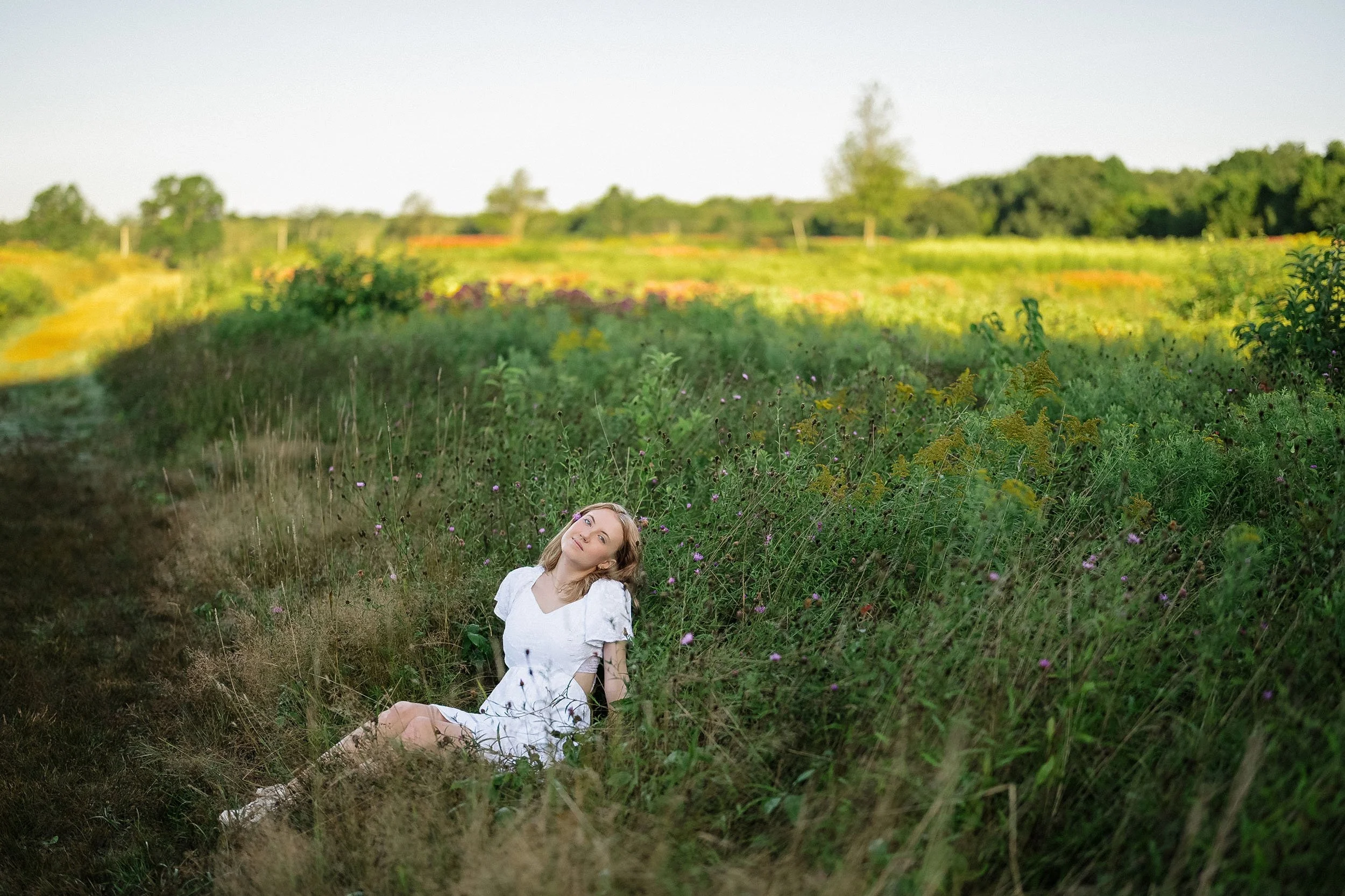 A woman in a white dress sitting in tall grass and wildflowers in a green field with trees and hills in the background under a clear sky.