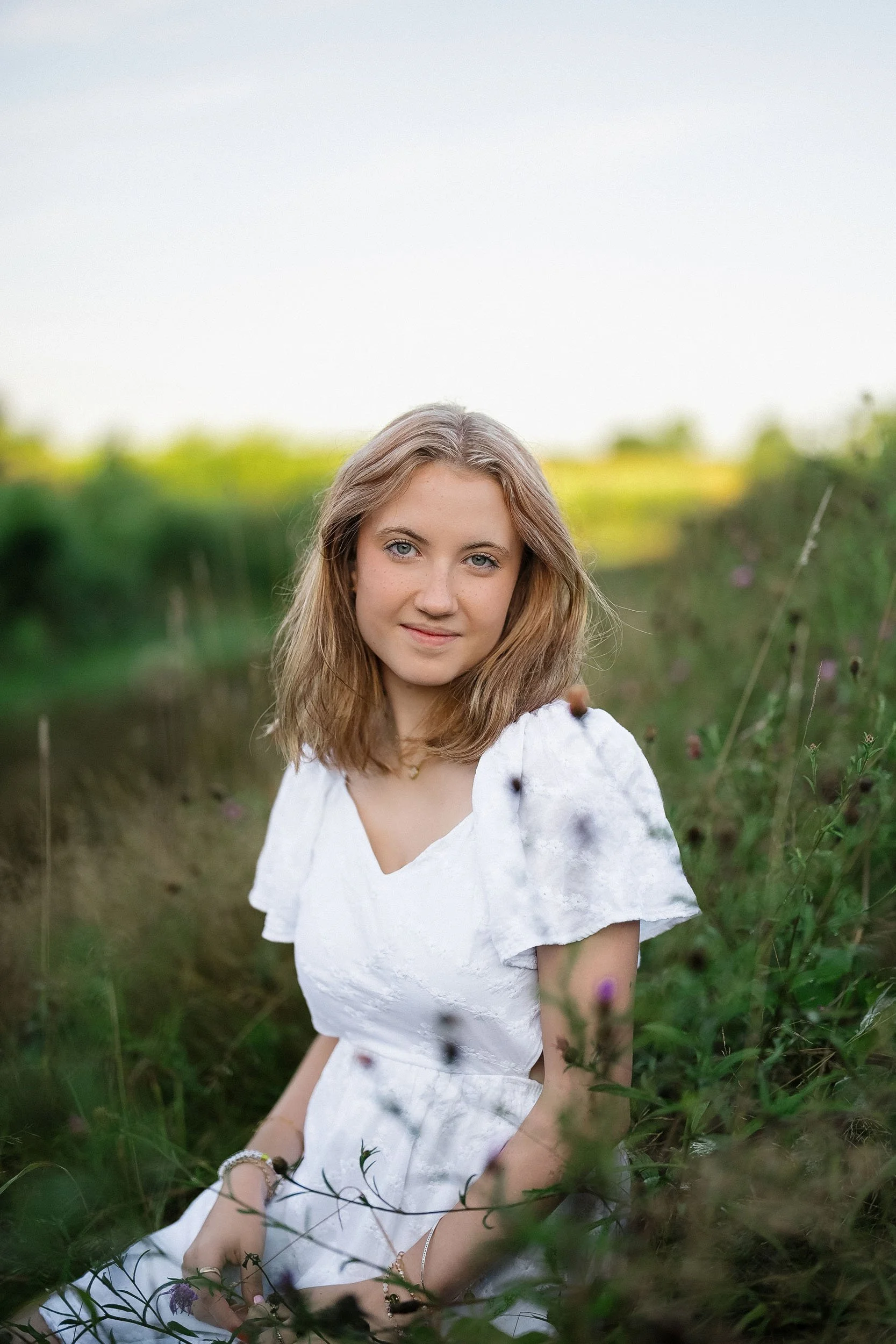 A young woman sitting in a grassy field with wildflowers, wearing a white dress with ruffled sleeves, and looking at the camera with a slight smile.