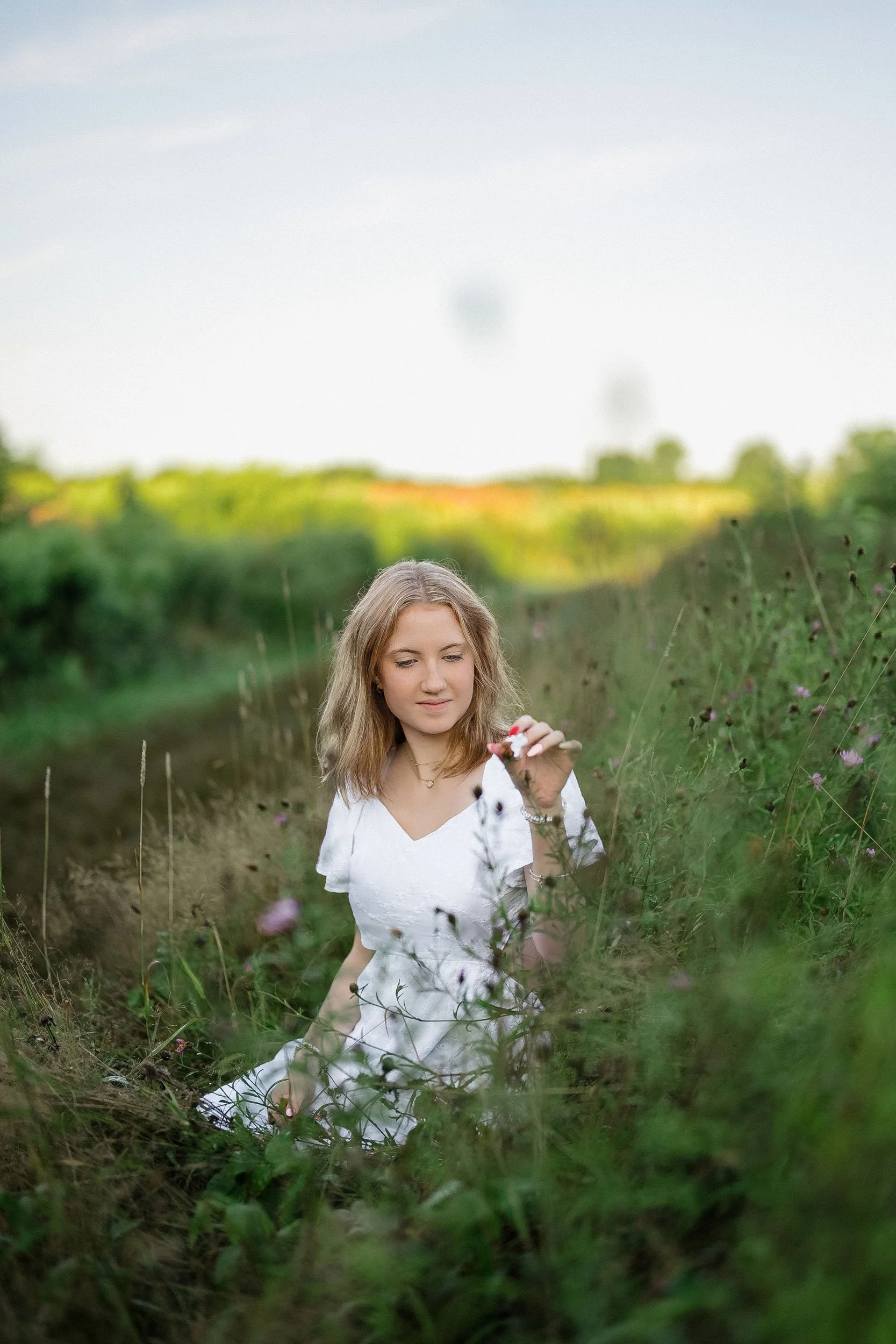 A young woman in a white dress kneeling in a grassy field, surrounded by wildflowers, holding a small flower and looking at it thoughtfully, with a blurred green landscape and a blue sky in the background.