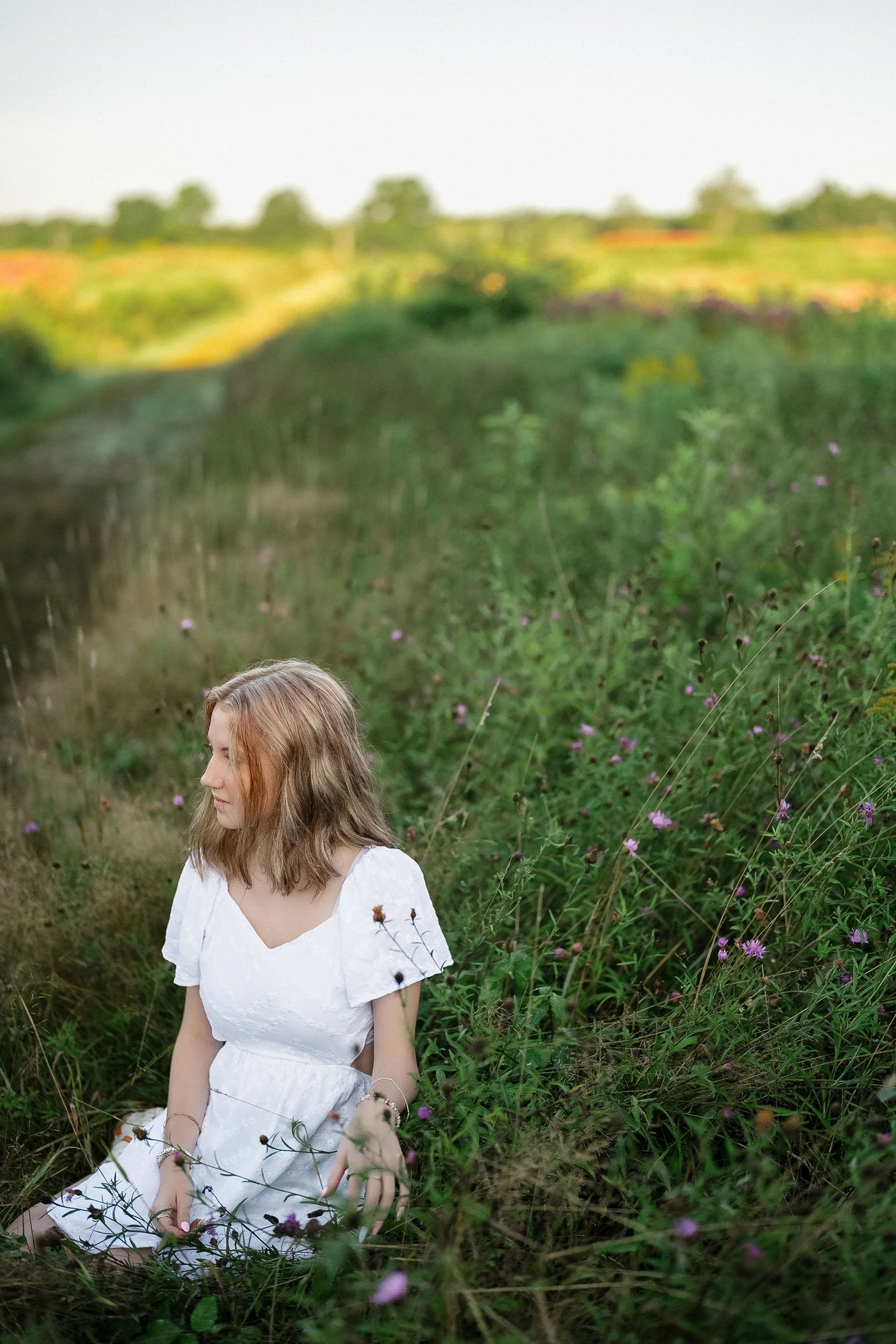 A young woman with light brown hair sitting on the grass next to pink flowers in a green field.
