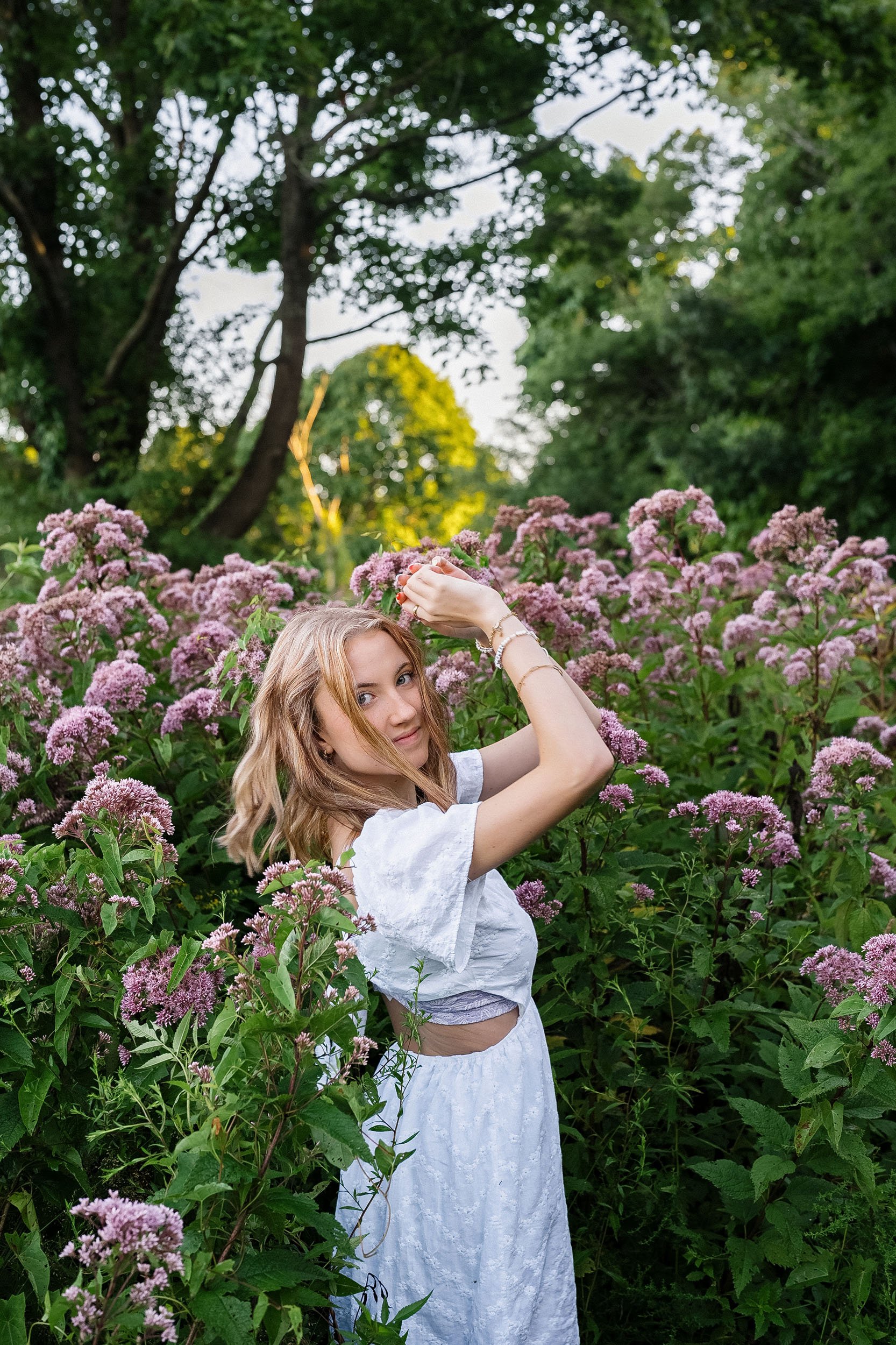 A young woman with blonde hair, wearing a white dress, standing among pink flowers in a lush green outdoor setting.