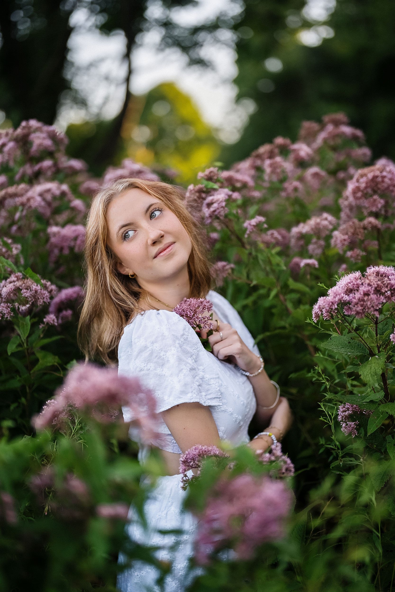A woman with shoulder-length light brown hair and blue eyes standing amidst pink flowering plants in a garden, wearing a white dress and holding a flower close to her chest.