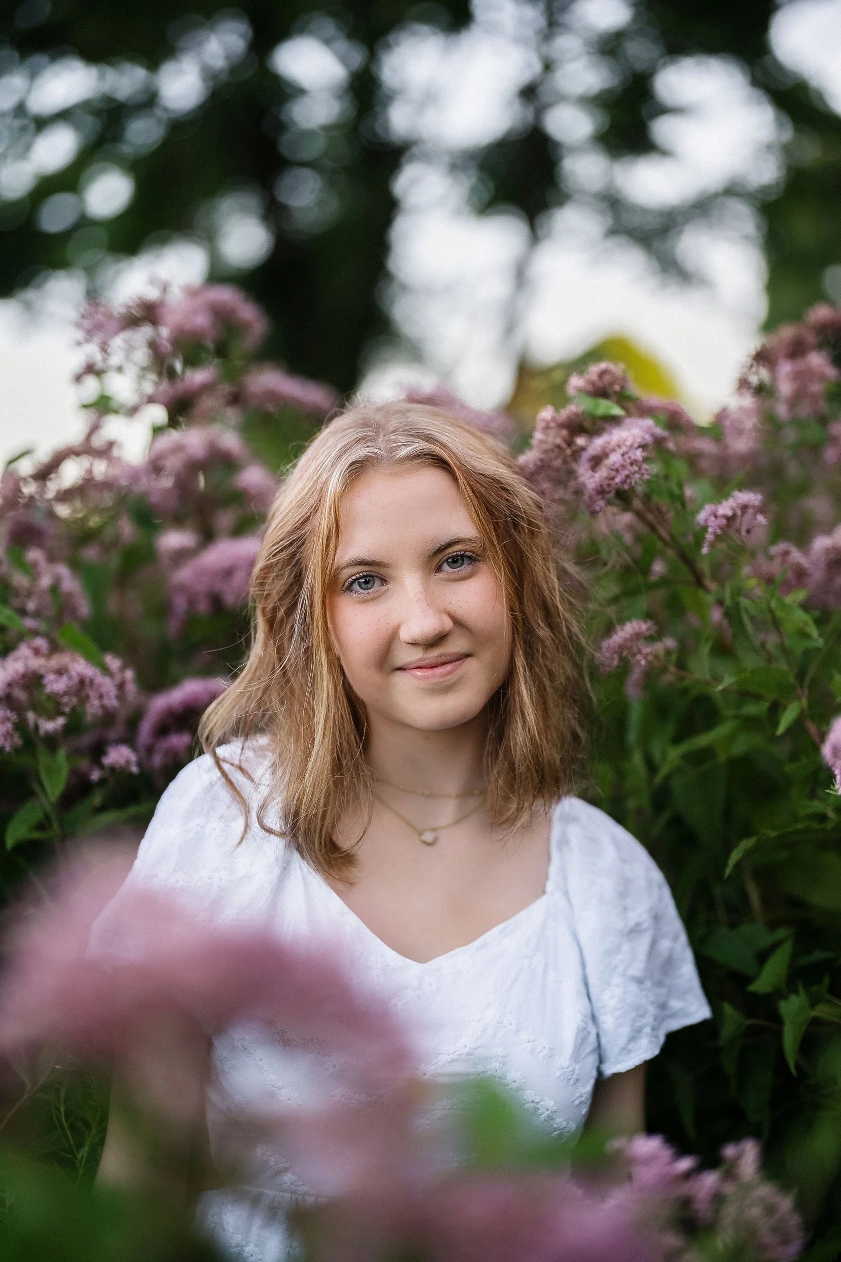 A young woman with light skin and wavy, shoulder-length blonde hair stands among pink flowers and green foliage, looking at the camera with a slight smile.