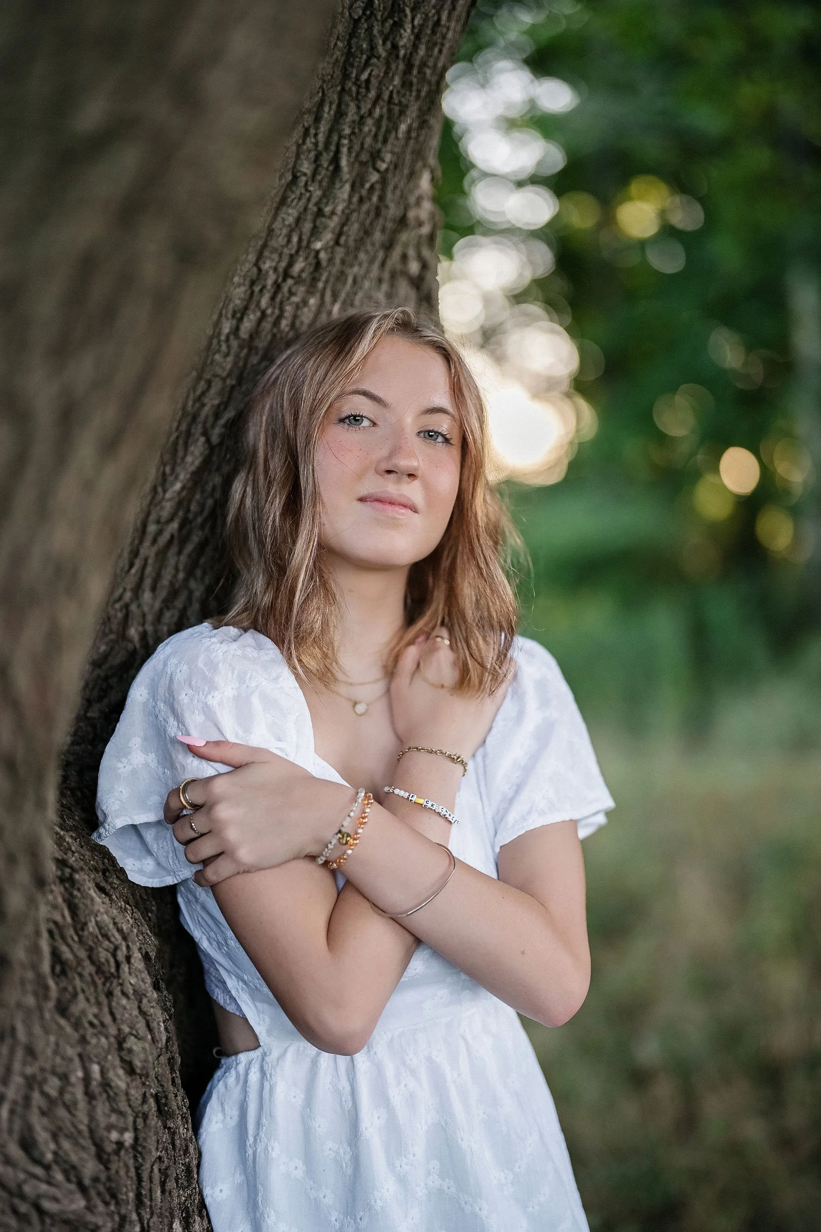 A young woman with light brown, shoulder-length hair leaning against a tree outdoors during daylight. She is wearing a white dress with puffed sleeves and has multiple bracelets, rings, and a necklace. The background is blurred with greenery.