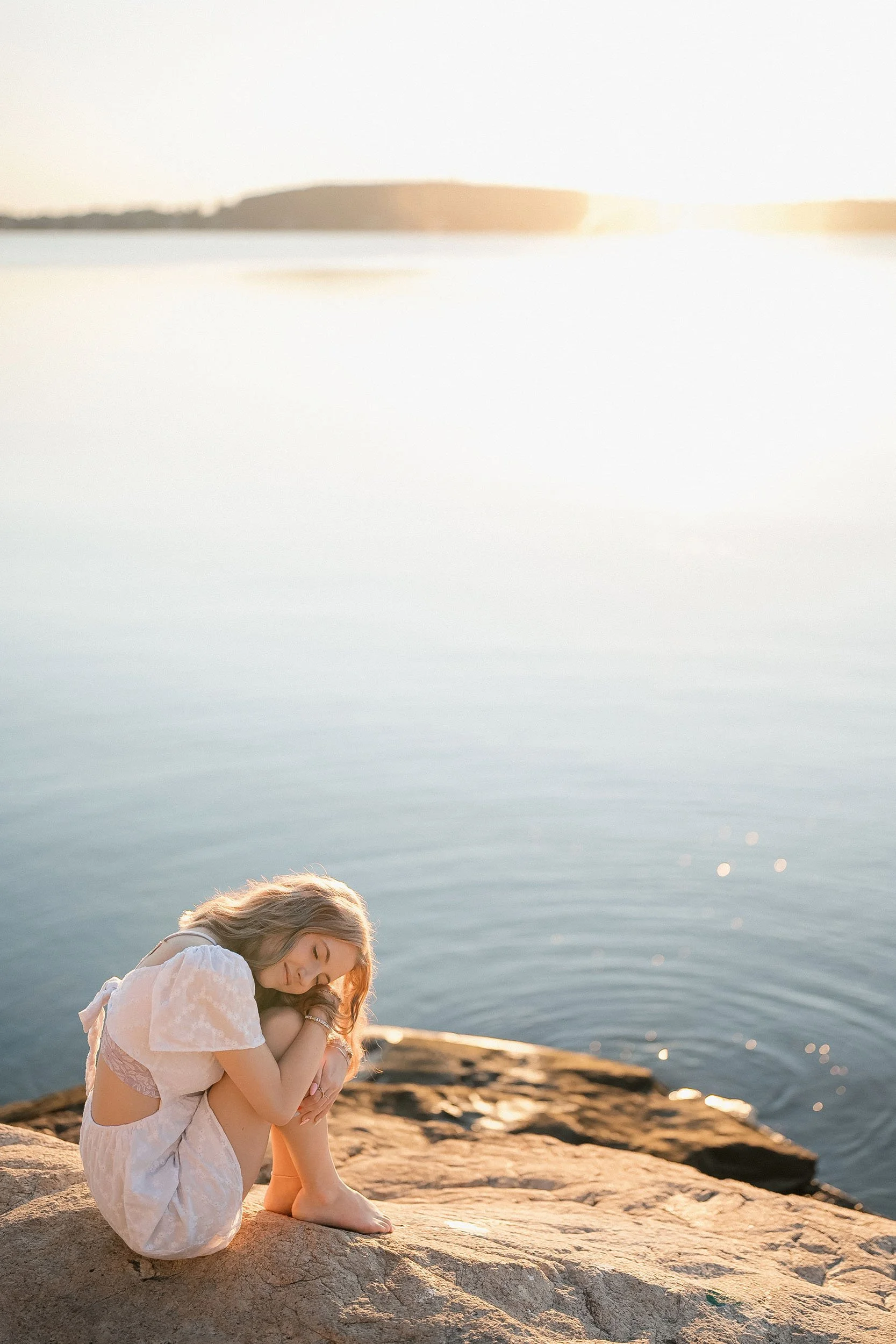 A woman sitting on a rock by the water with her head resting on her knees at sunset.