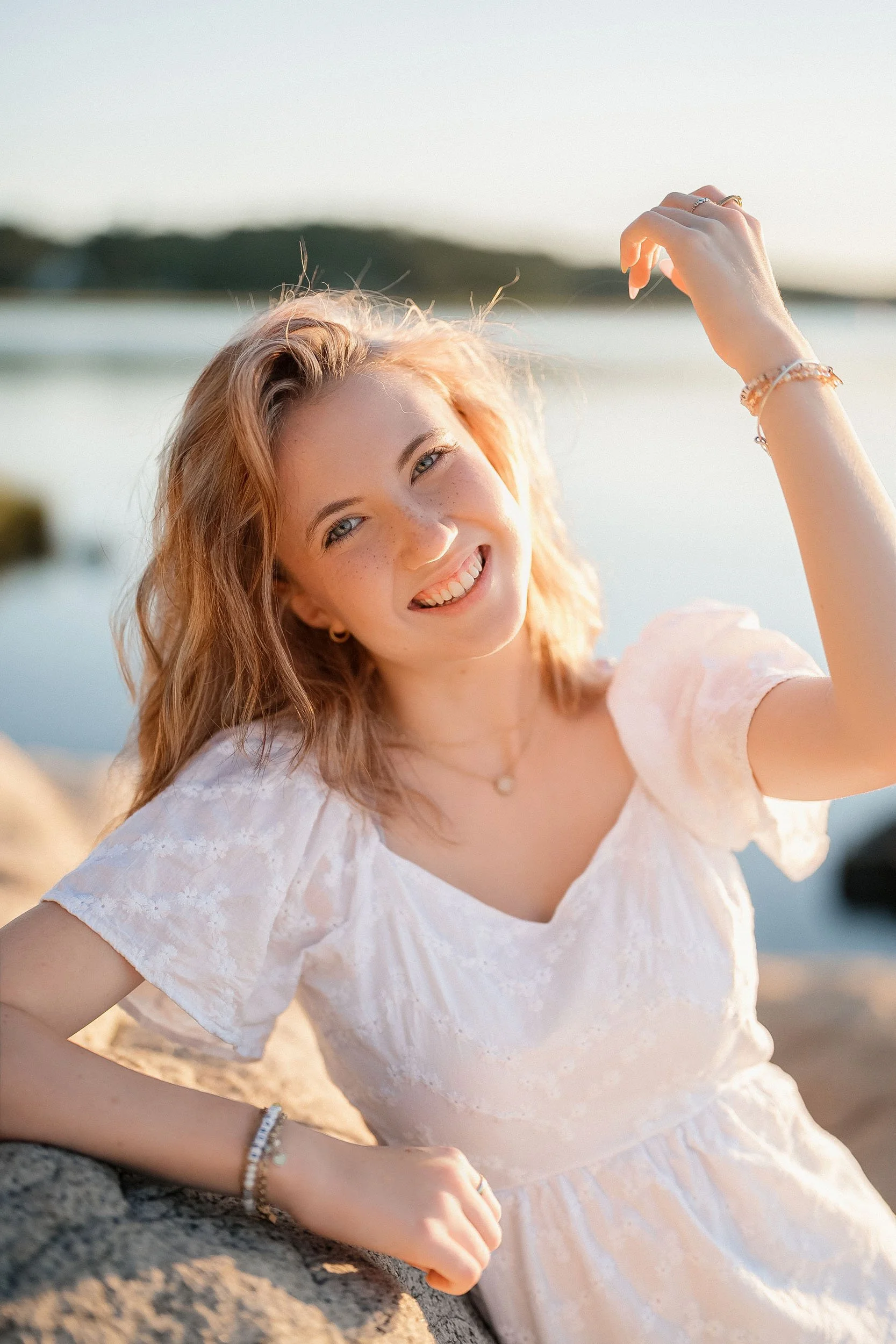 A young woman with curly blonde hair and freckles smiling and looking at the camera while sitting on rocks by a body of water during sunset.