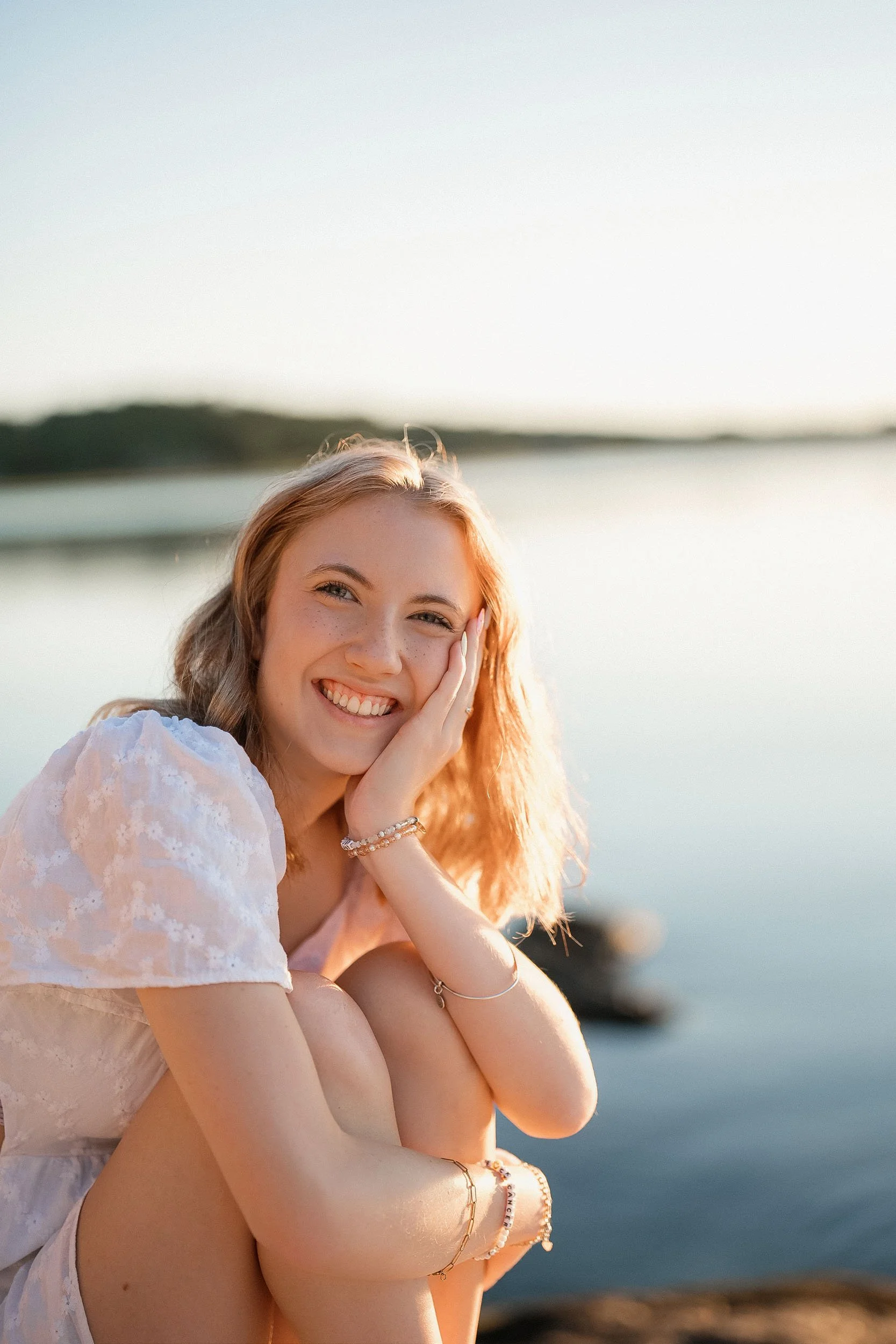 A young woman with red hair and freckles smiling by a body of water at sunset, sitting with her knees up and hand on her face, wearing a light-colored dress with jewelry.