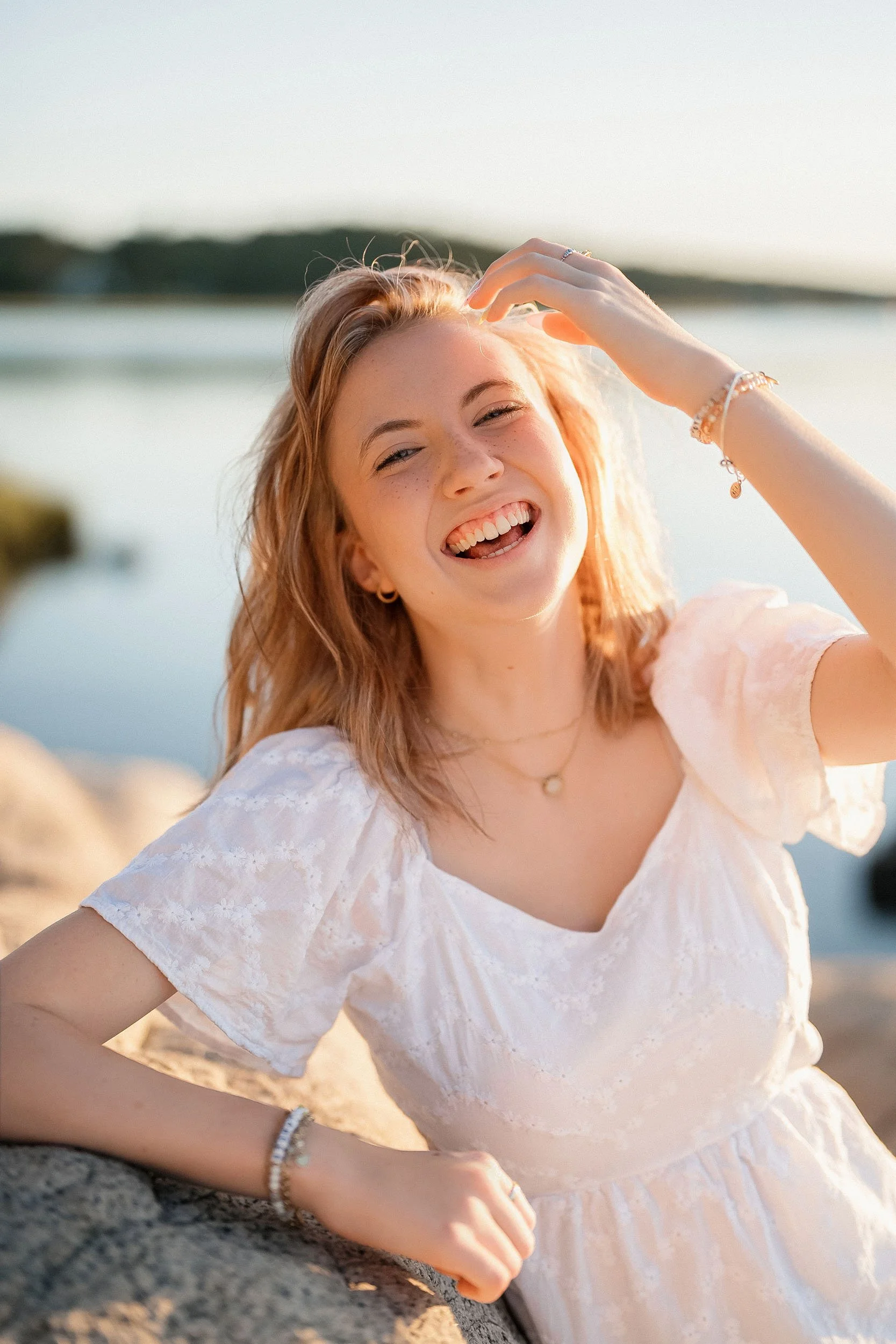 A young woman with red hair smiling and laughing outdoors near water with trees in the background, wearing a white dress and jewelry.