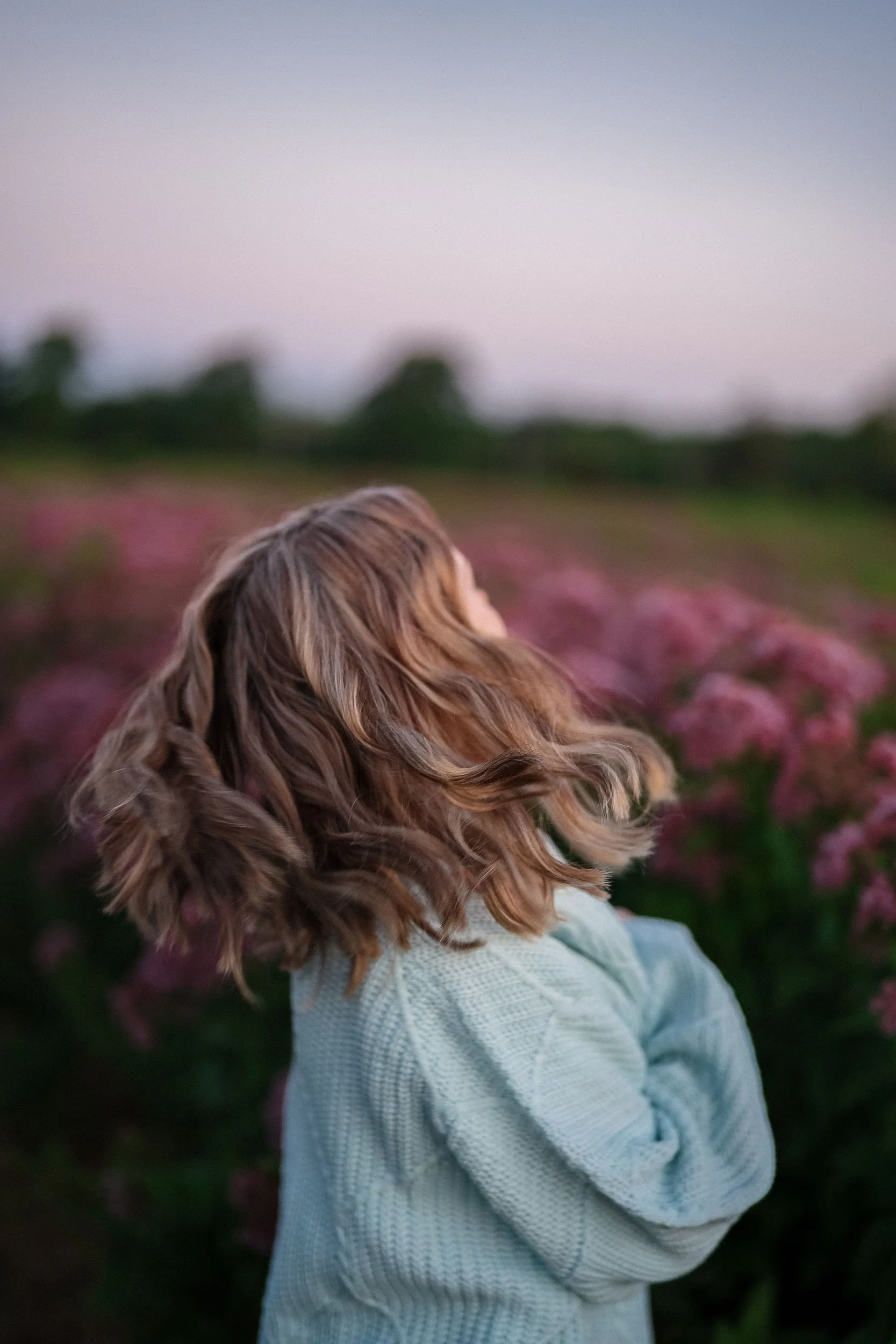 A woman with wavy, shoulder-length hair wearing a light-colored sweater standing in a field of pink flowers during sunset.