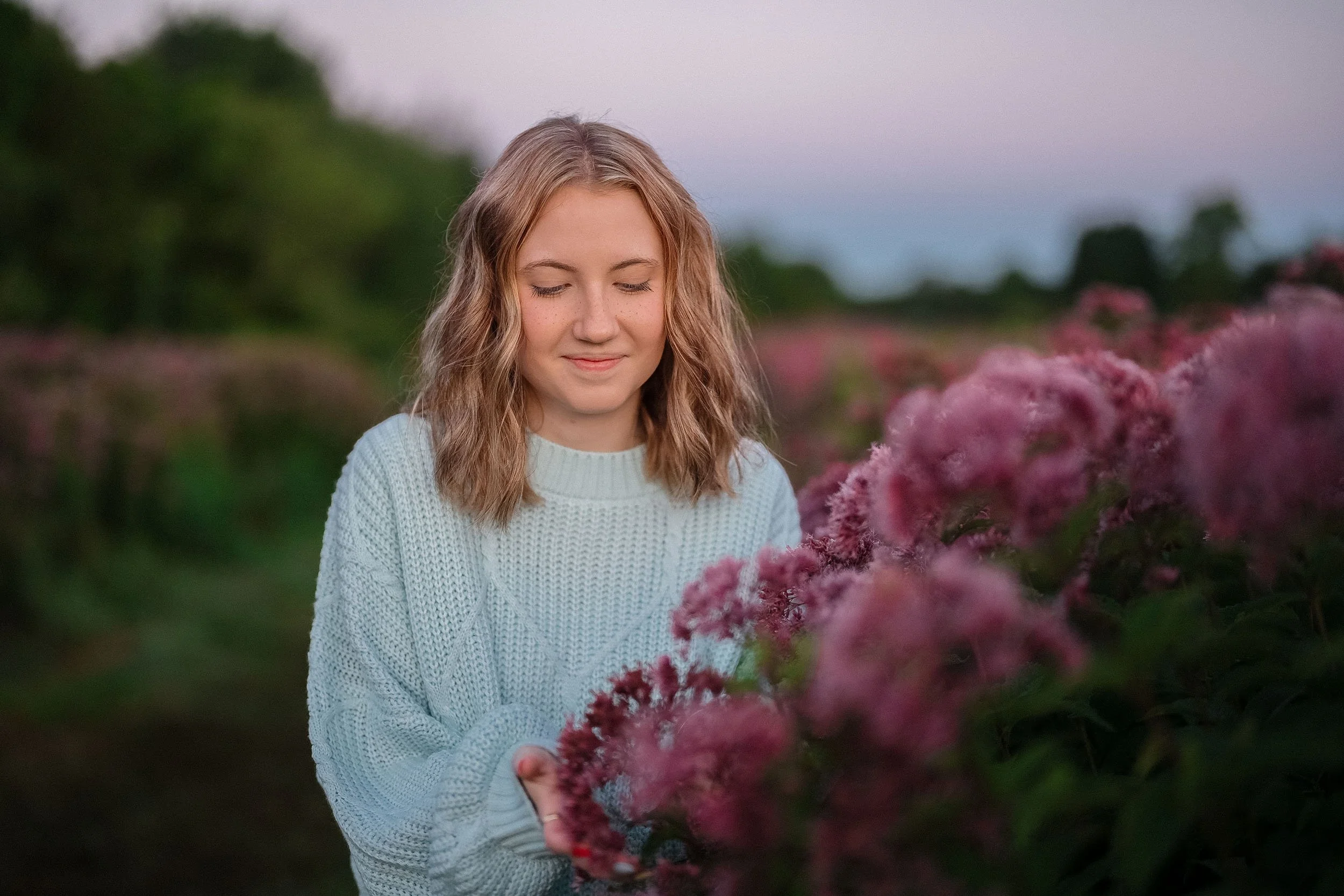 A young woman with wavy blonde hair wearing a light blue sweater, standing outdoors among pink flowers with her eyes closed and a gentle smile.