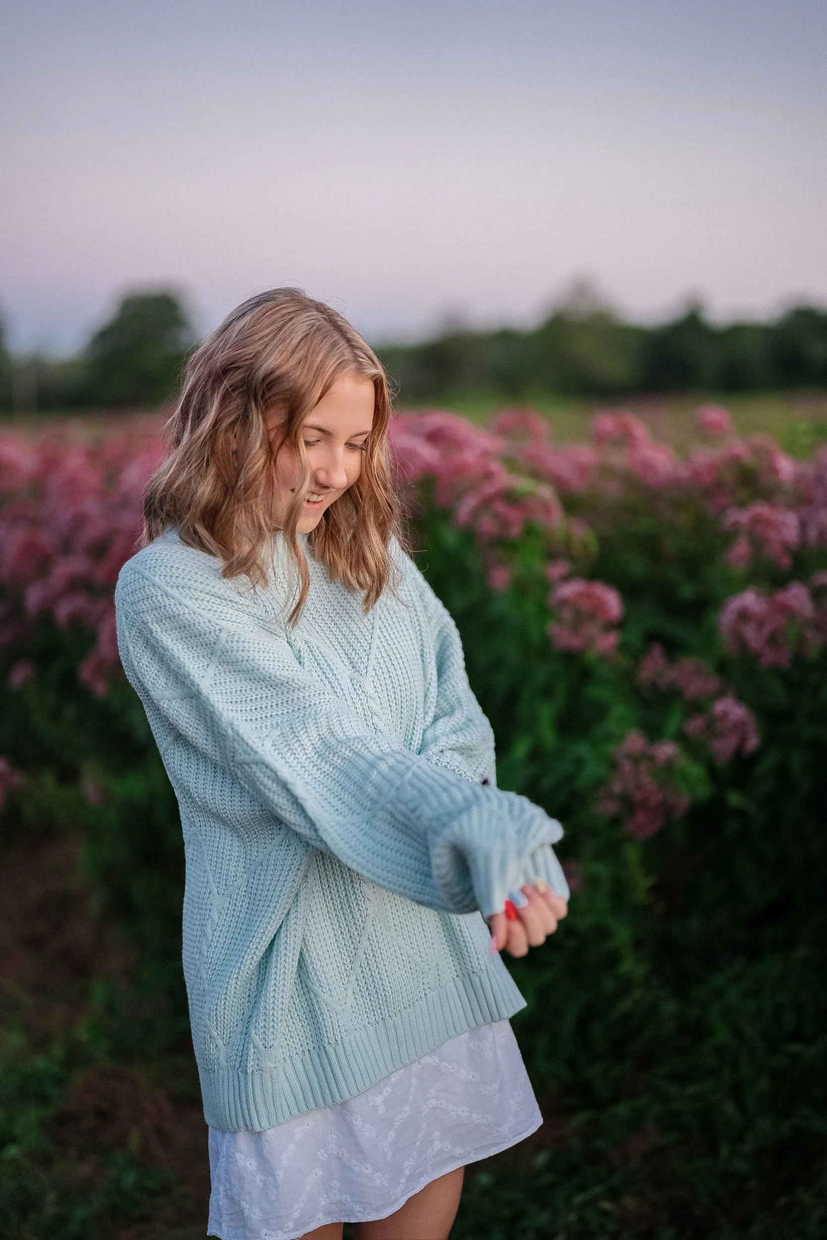 A young girl with wavy blonde hair wearing a light blue sweater and white skirt, smiling and looking down while standing in a field of pink flowers at dusk.