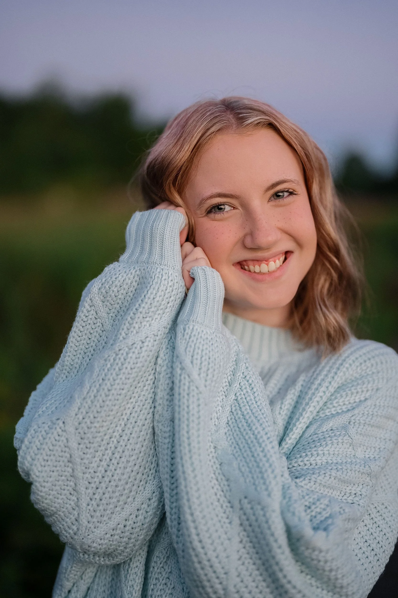 A young woman with shoulder-length red hair and freckles, smiling and wearing a light gray knit sweater, outdoors during dusk or dawn.