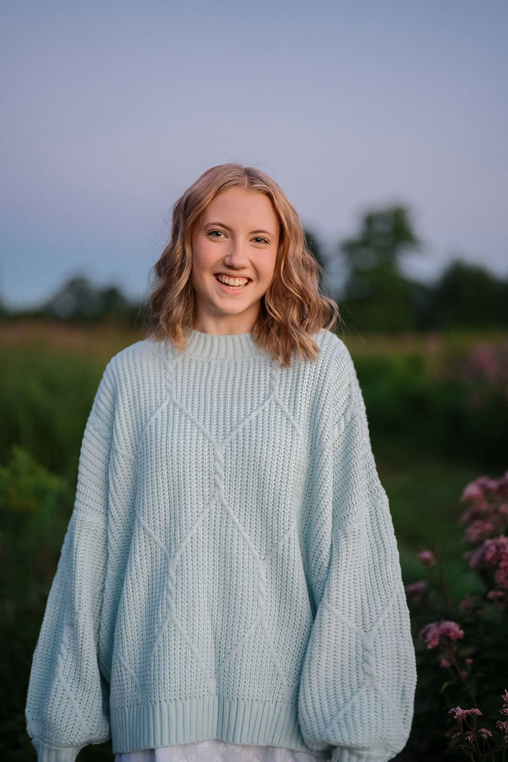 A young woman with curly blonde hair smiling outdoors during sunset, wearing a light gray cable-knit sweater in a green landscape.