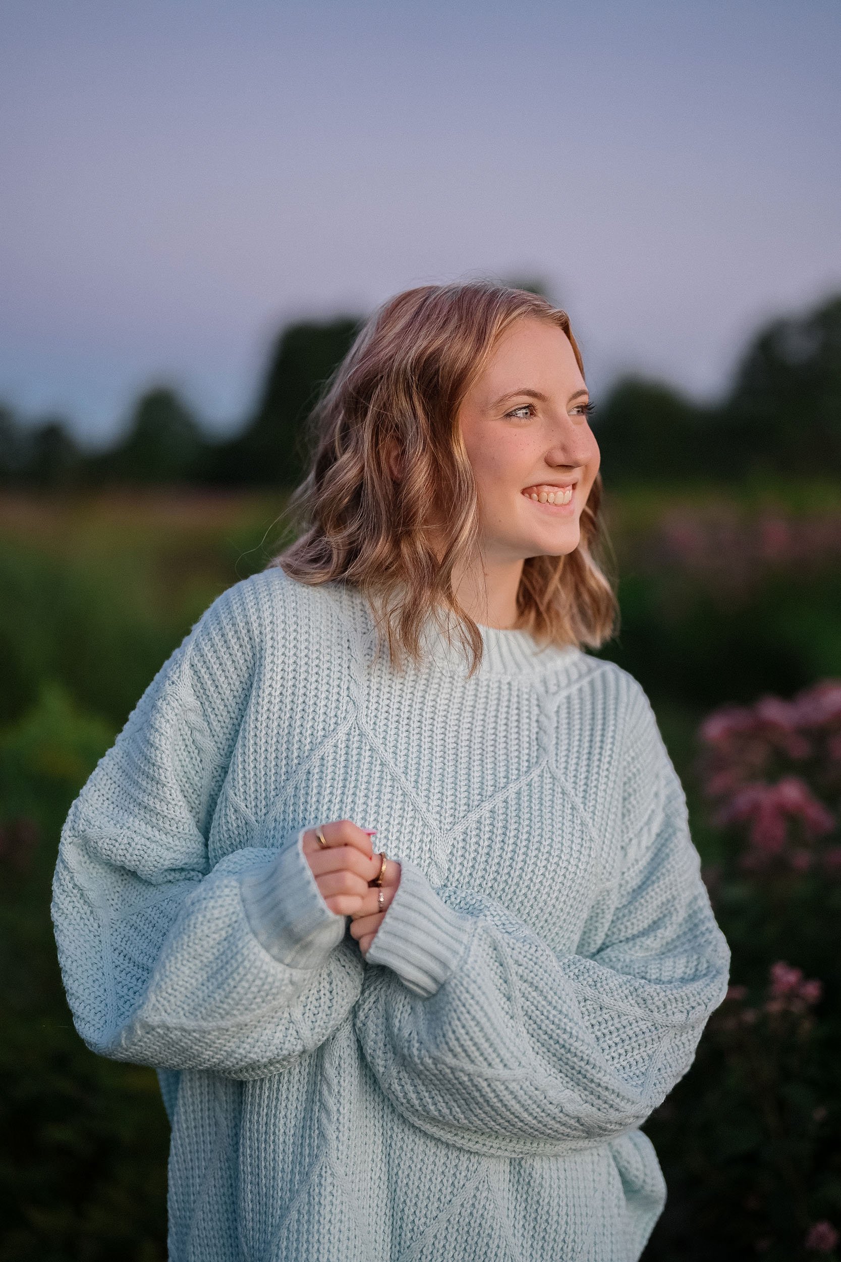 A young woman with wavy reddish-brown hair smiling while outdoors during dusk, wearing a cozy light blue knitted sweater, standing among green foliage and pink flowers.