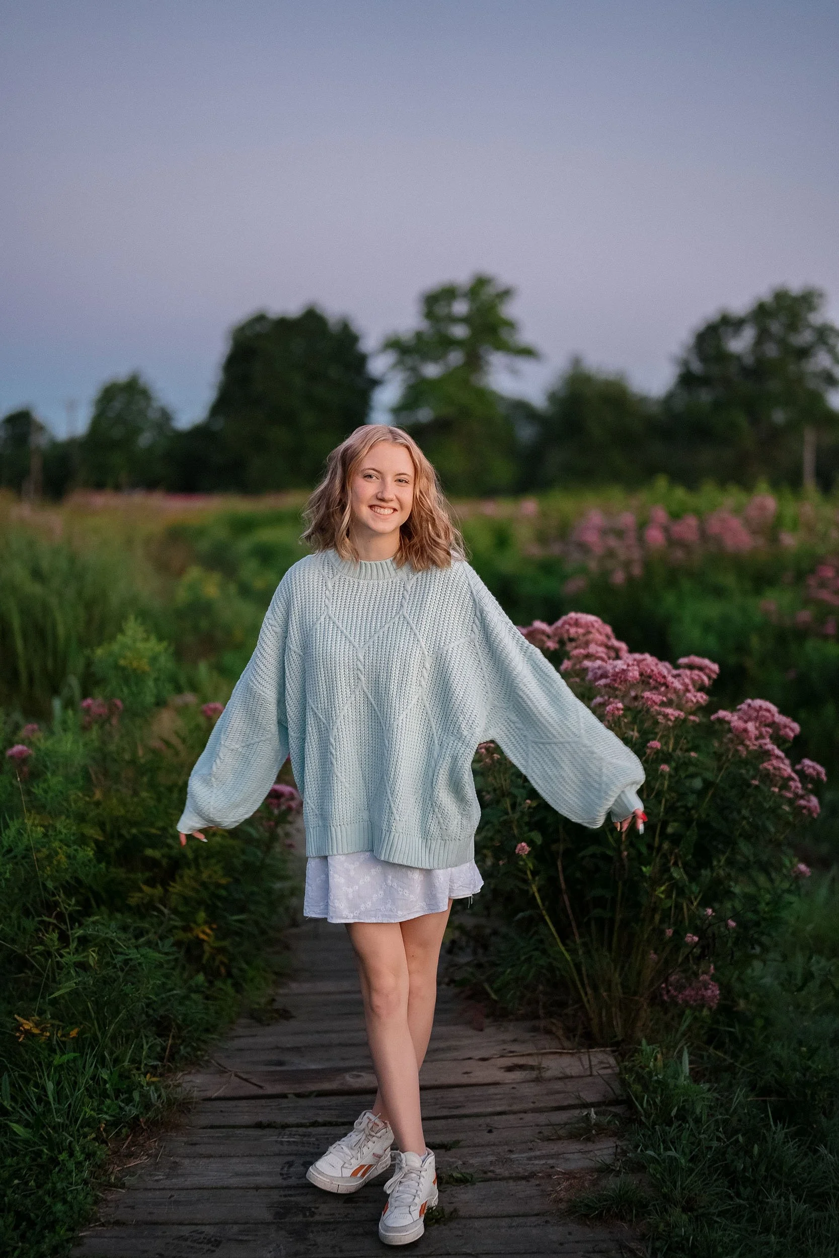 A young woman smiling and walking on a wooden pathway through a flower field at sunset, wearing a light blue oversized sweater, a white dress, and sneakers.
