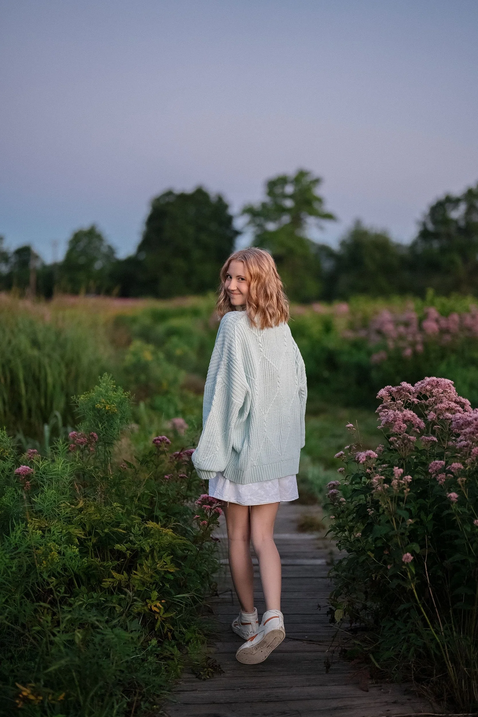 A young woman walking on a wooden path in a garden with pink flowers, wearing a light gray sweater, a white dress, and sneakers, looking back and smiling.