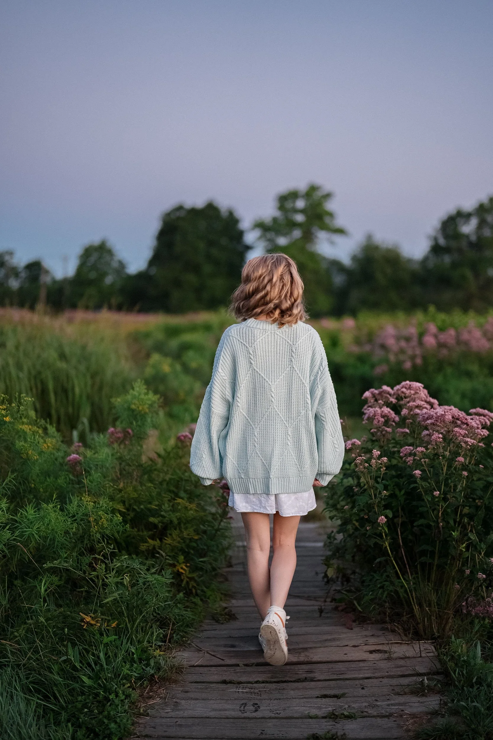 A girl with wavy, shoulder-length hair walking on a wooden pathway through a lush garden with pink and yellow flowers, with green trees in the background at dusk.