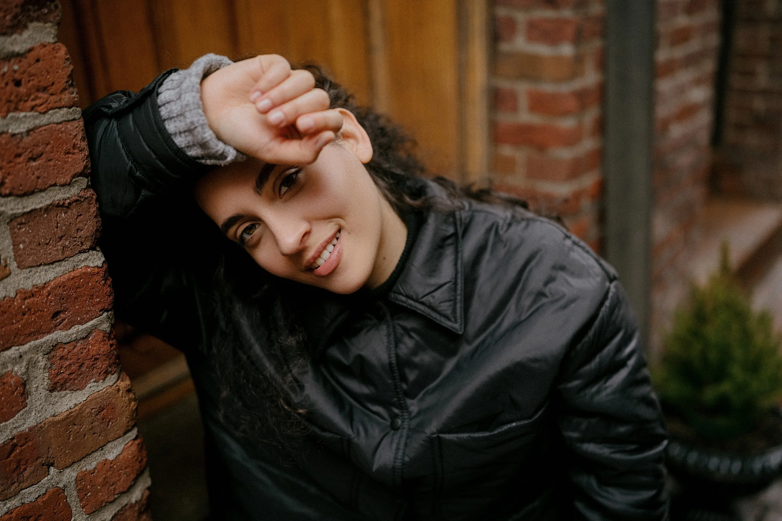 Young woman with curly dark hair smiling, leaning against a brick wall, wearing a black jacket and a gray sweater.