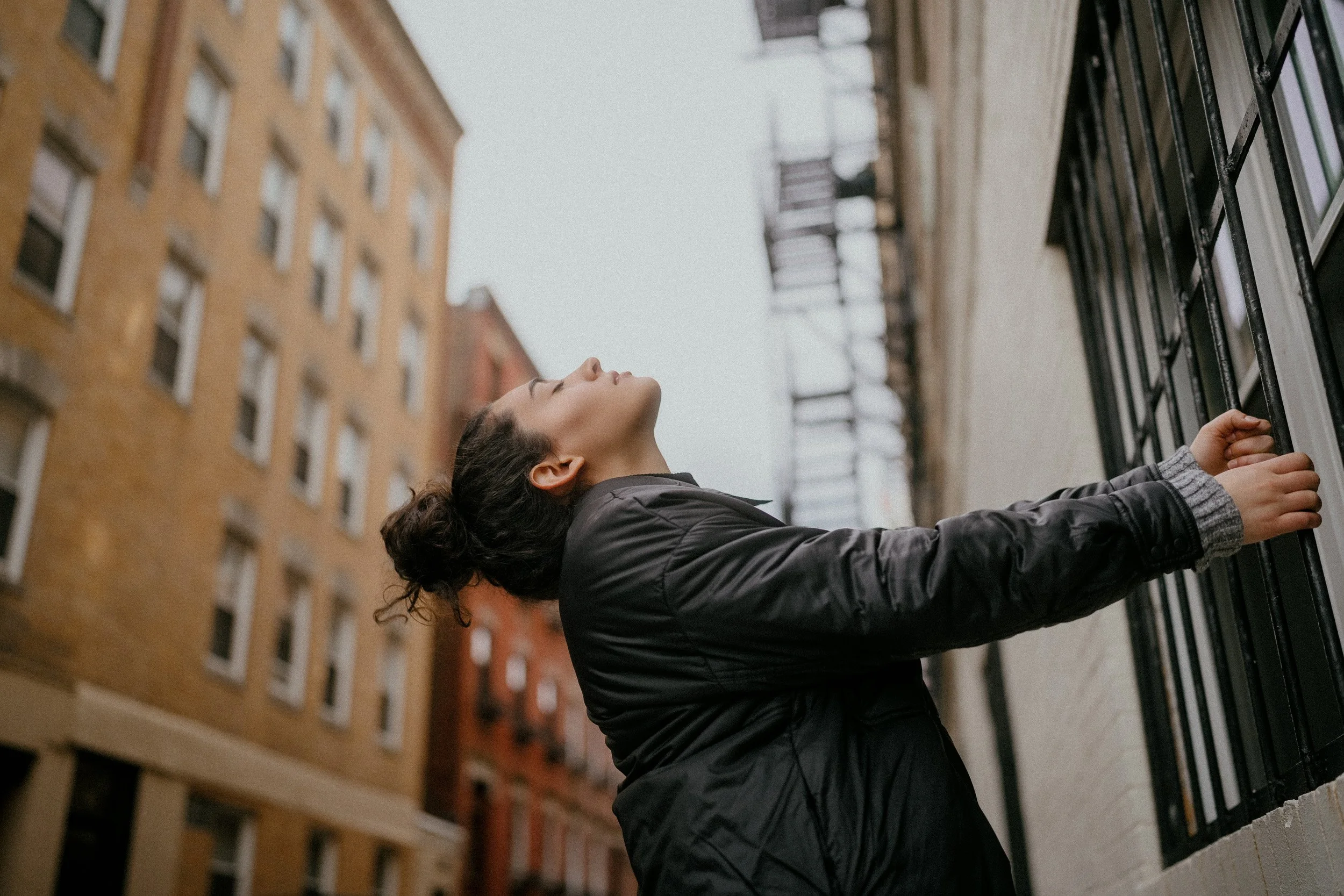 A woman with dark hair in a bun wearing a black jacket, reaching out towards a window with black bars on an urban building.
