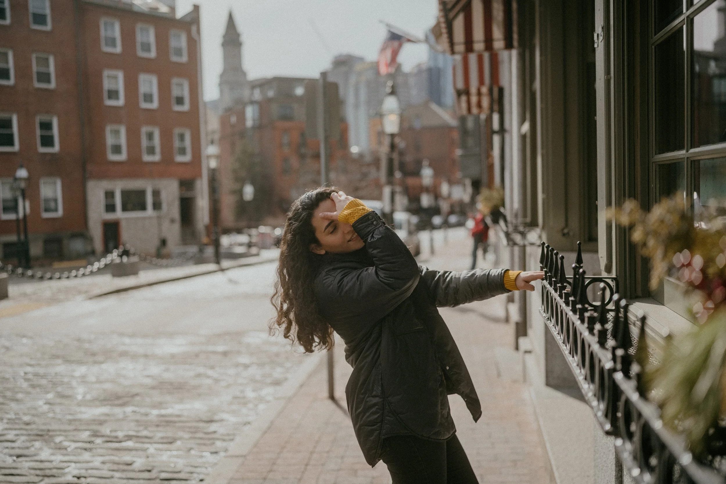 A woman with curly hair wearing a black coat and yellow sweater, standing on a city sidewalk, covering her face with one hand and reaching out with the other hand towards a window with a black decorative railing.