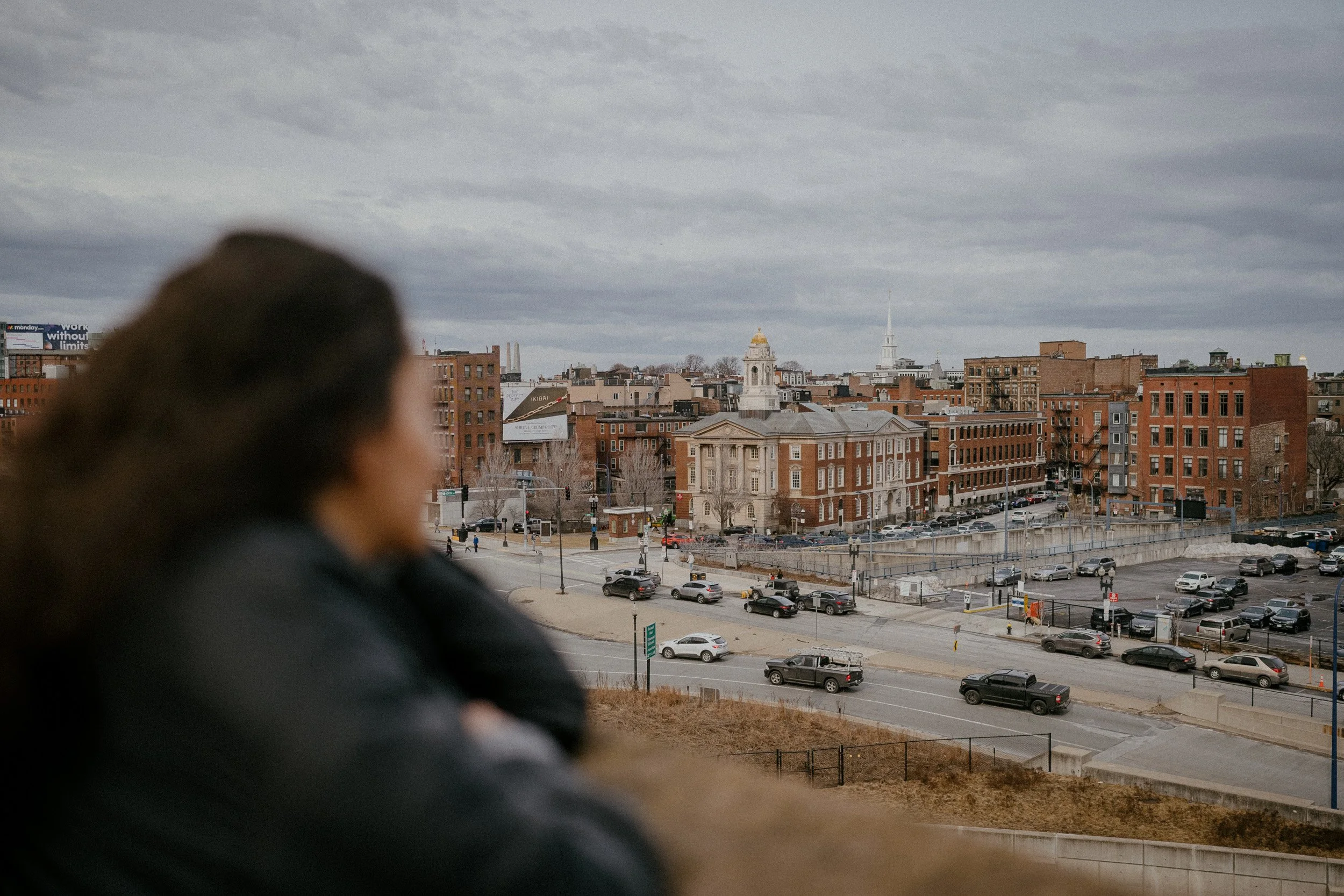 Blurred person sitting on a ledge overlooking a cityscape with brick buildings, parking lots, and a cloudy sky.