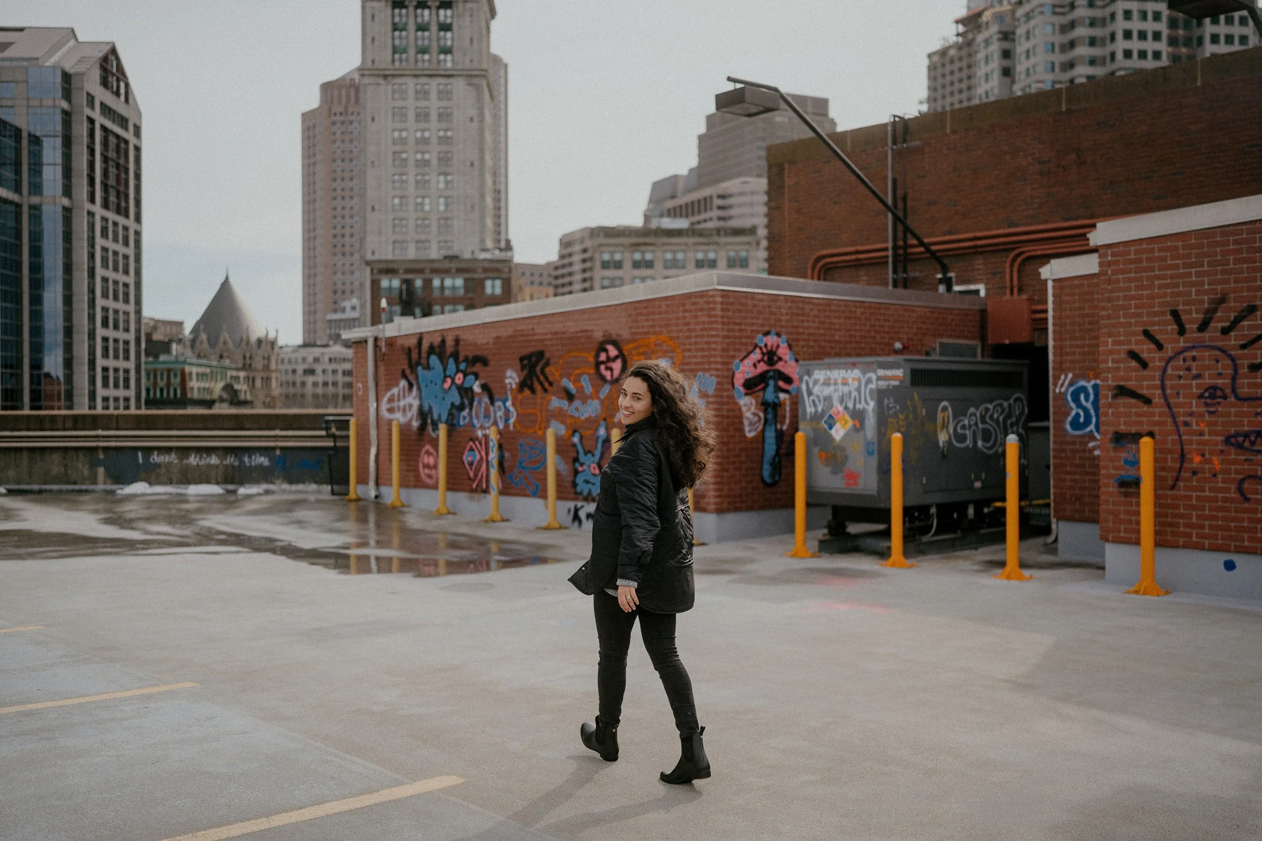 A woman with curly hair in a black jacket and black boots walking on a city rooftop parking lot with graffiti on brick walls and tall buildings in the background.