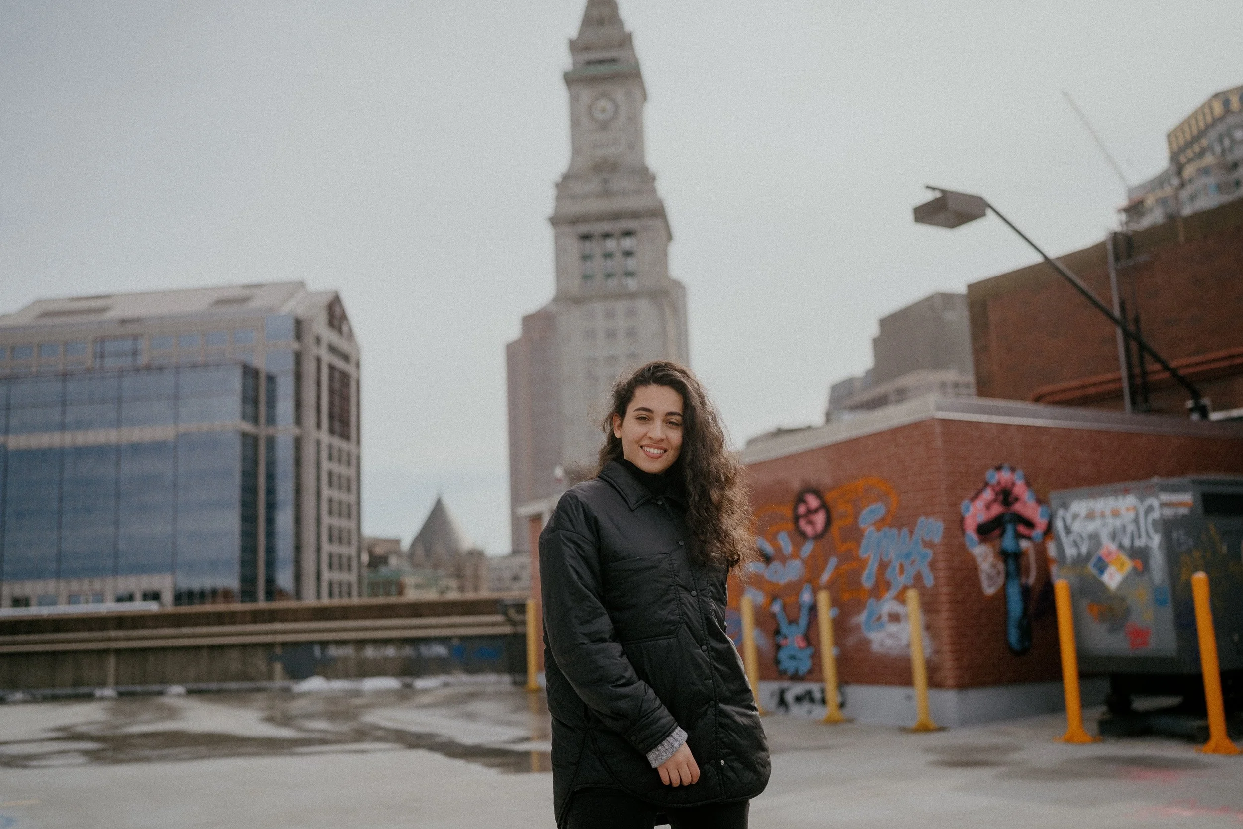 A young woman with long curly hair smiling and standing in an urban setting with city buildings and a clock tower in the background.