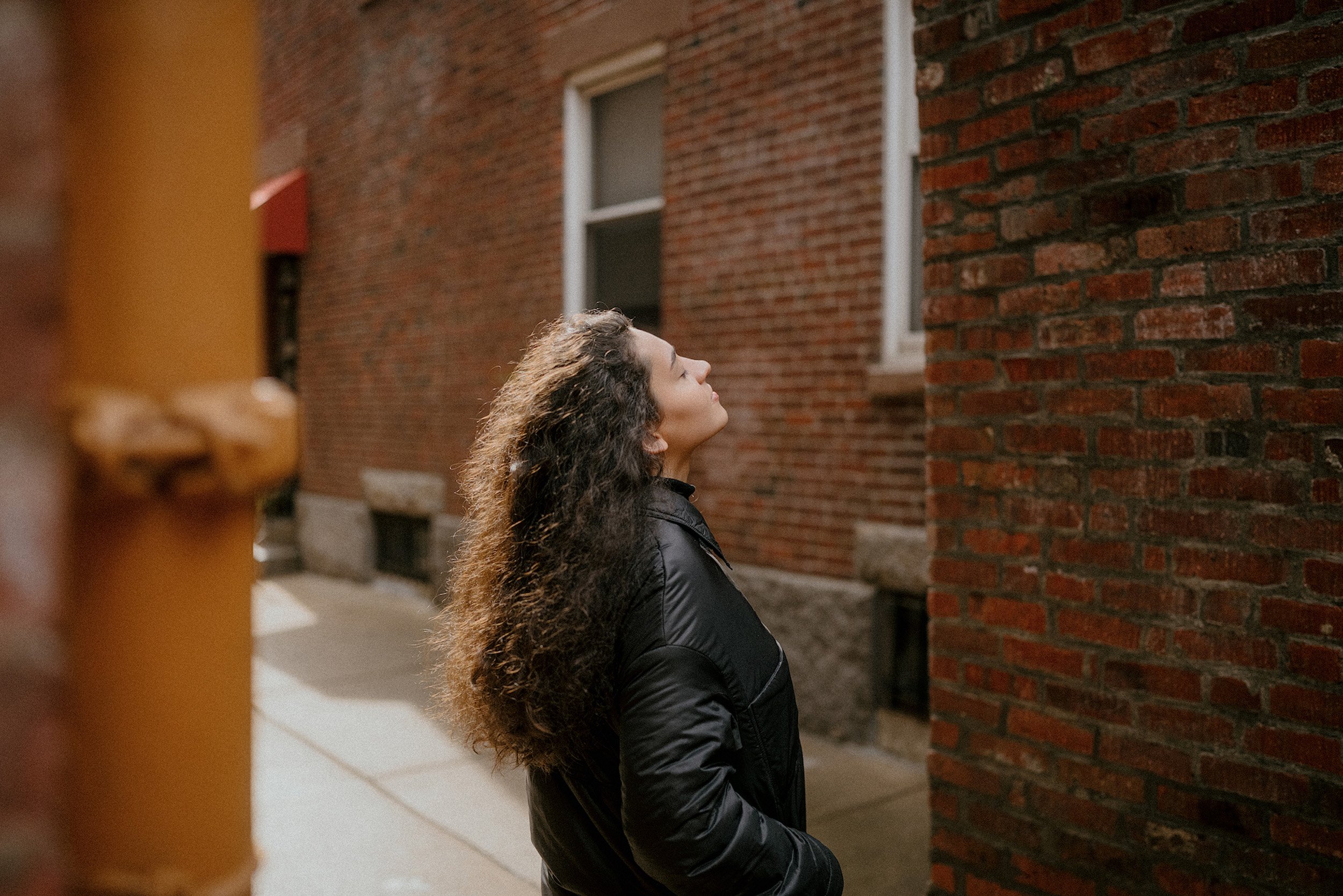 A woman with long, curly hair wearing a black jacket standing on a sidewalk between brick buildings, looking upward with a serene expression.