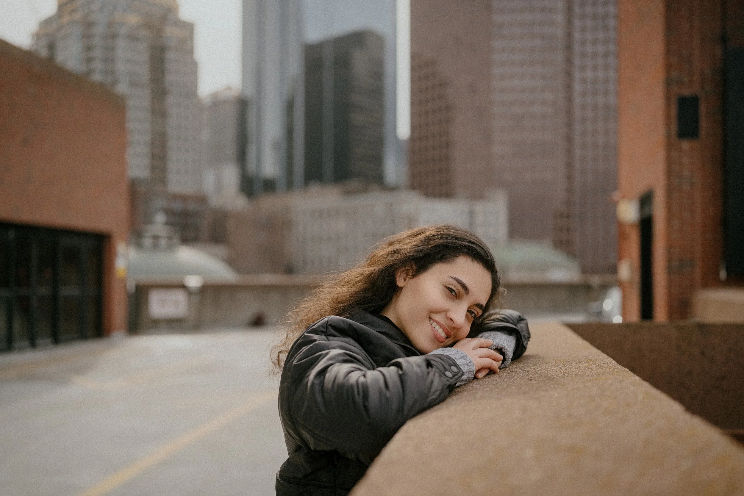 Smiling young woman leaning on a rooftop ledge with city skyscrapers in the background.