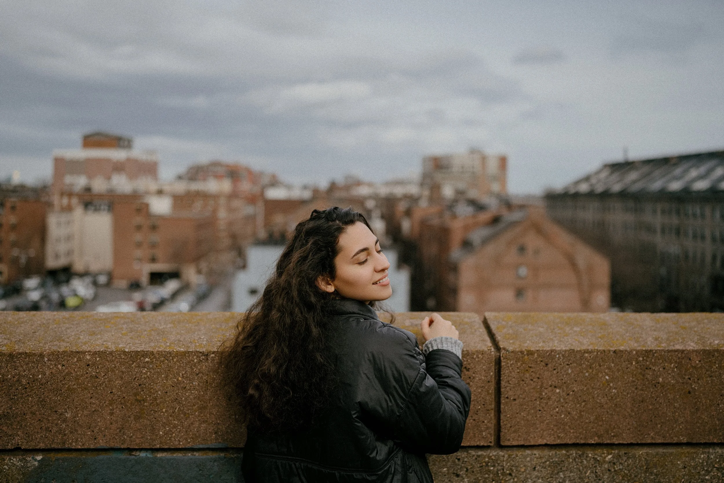 Young woman with curly hair smiling with eyes closed, wearing a black jacket, standing on a rooftop with an urban cityscape in the background.