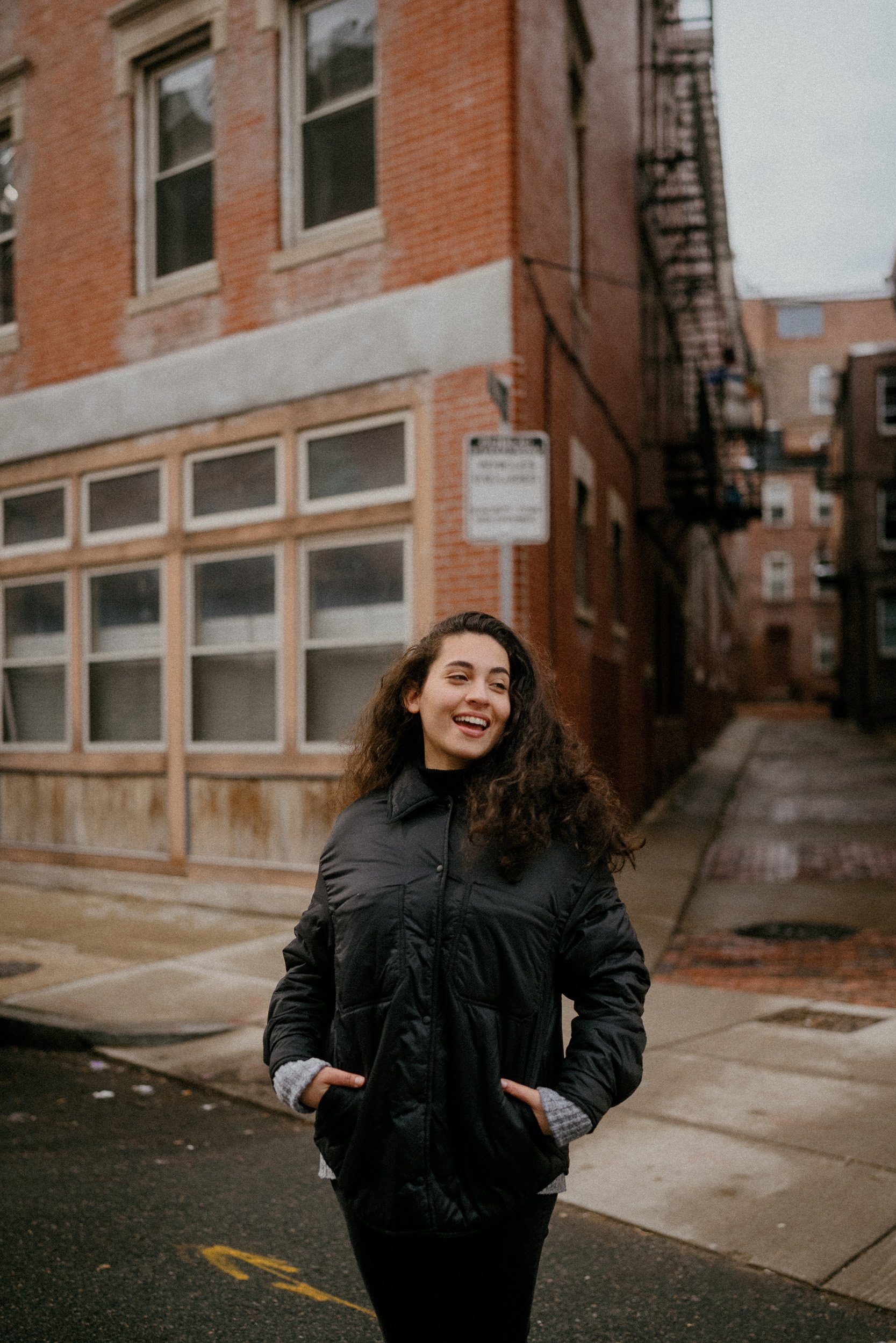 A young woman with curly hair, smiling, wearing a black jacket, walking on an urban street with brick buildings in the background.