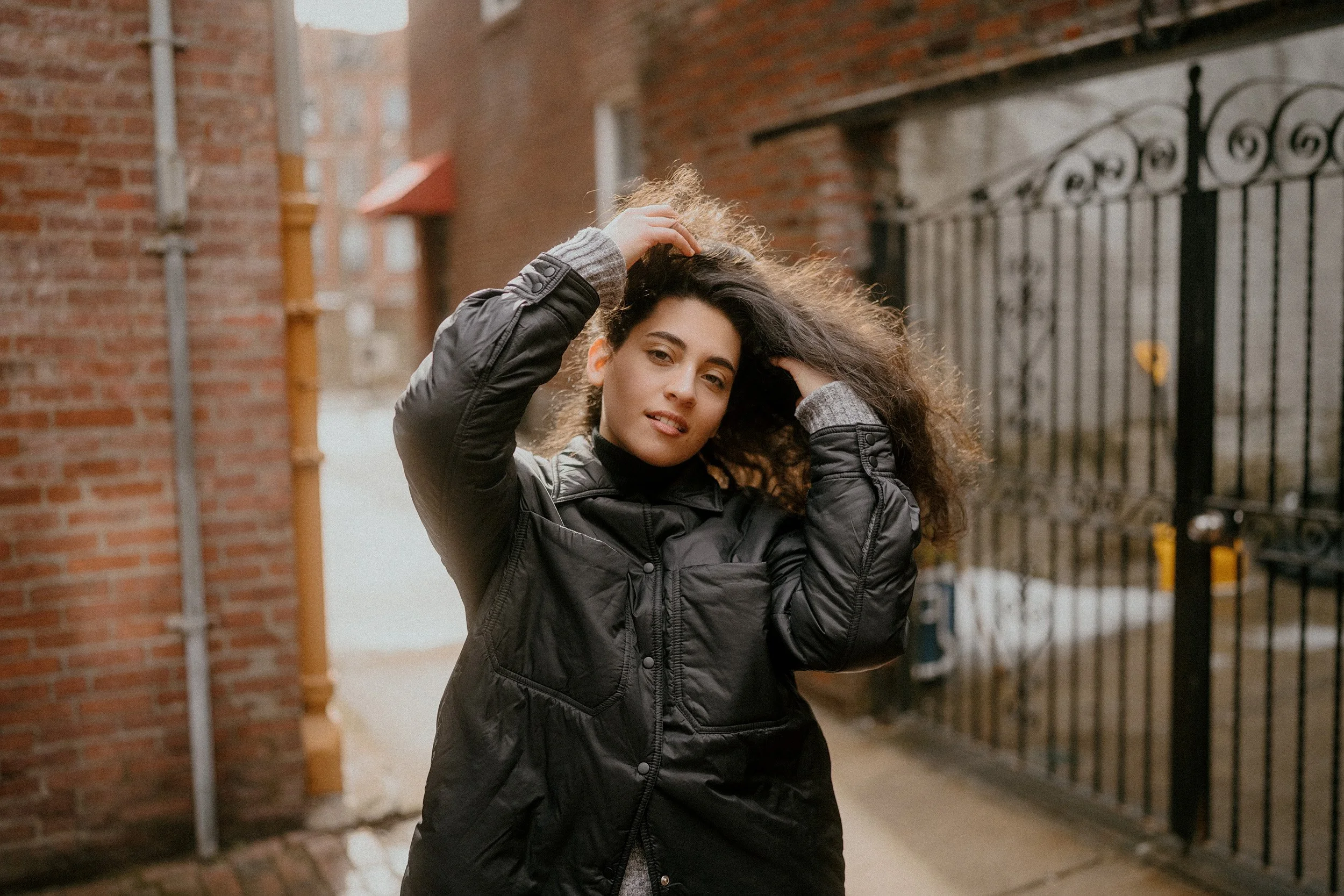 A woman with curly hair standing outdoors in an alley, touching her hair and looking at the camera, wearing a black jacket and gray sweater.