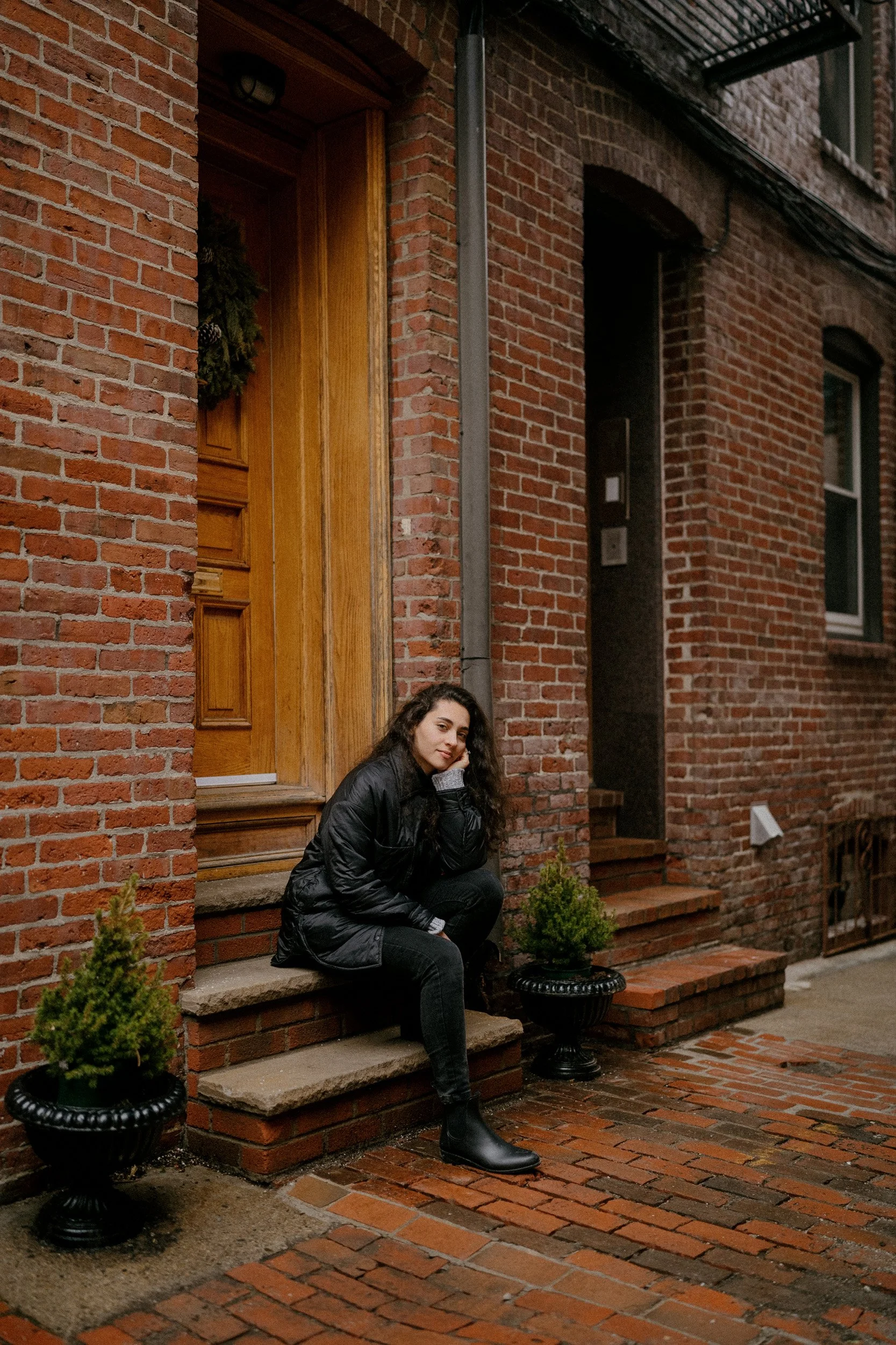 A young woman in black clothing and boots sitting on brick steps outside a brick building with a wooden door, flanked by two potted small evergreen trees.